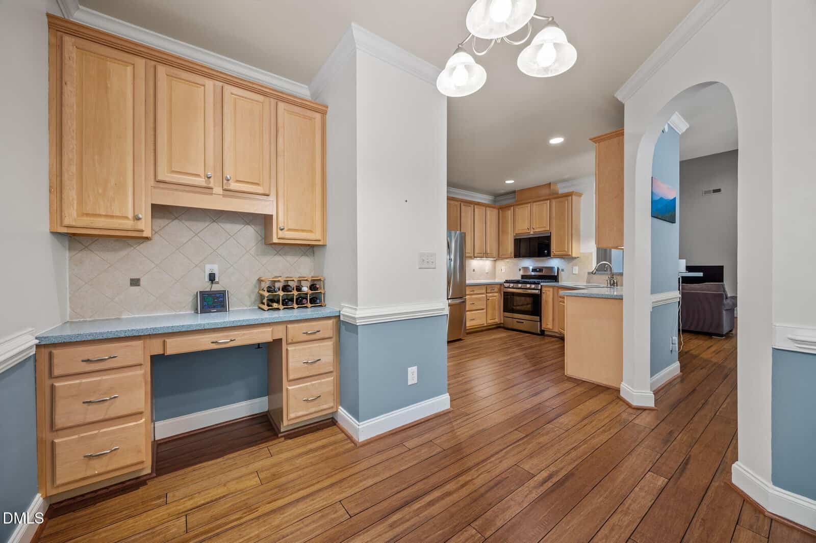 A kitchen with light wood cabinets, stainless steel appliances, wood flooring, and a built-in desk area. An arched doorway leads to a living room with a sofa and a wall-mounted TV.
