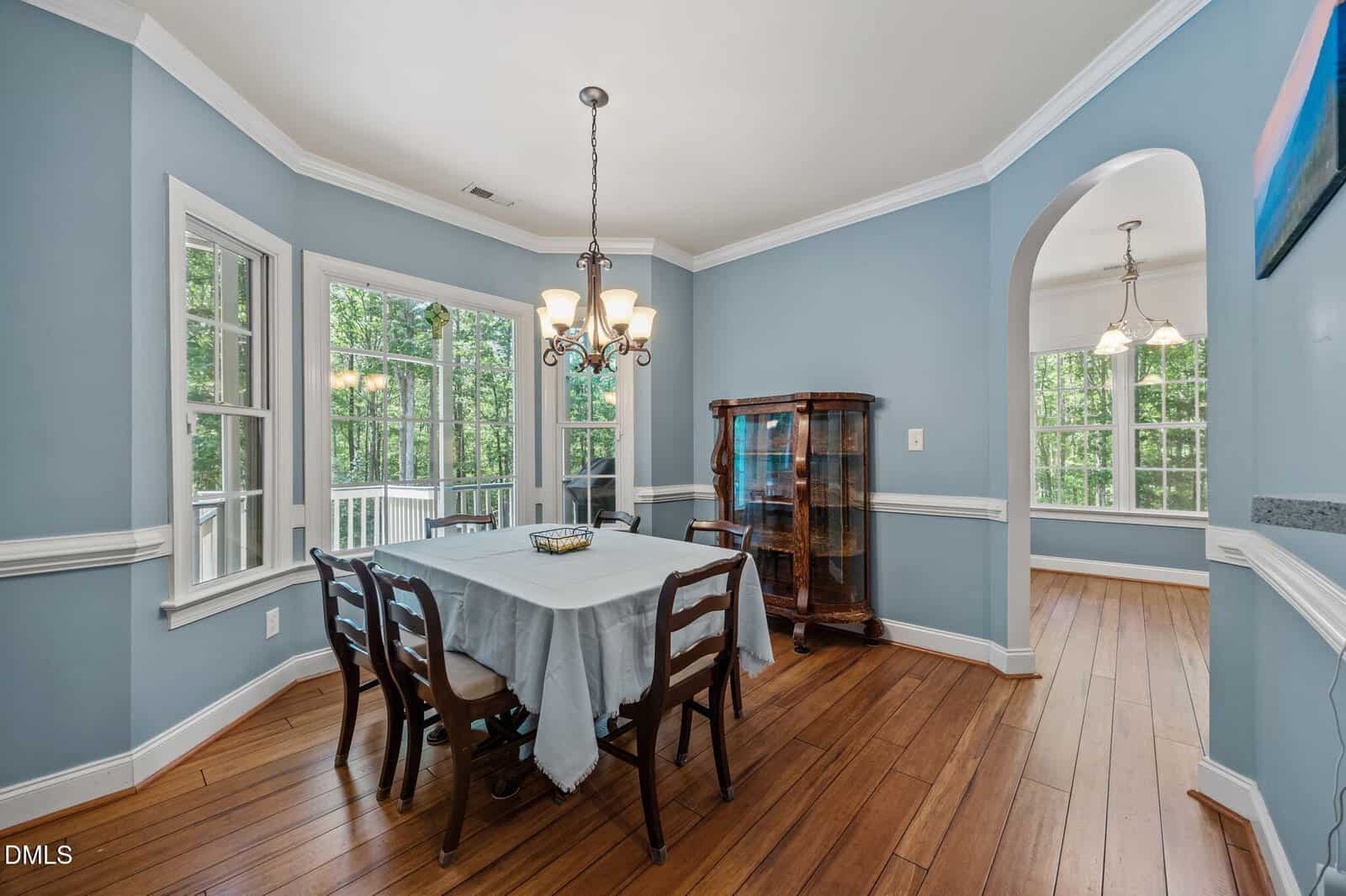 A blue dining room with a wooden table and six chairs, chandelier above, large windows showing greenery outside, wood floors, and an arched doorway leading to another room.