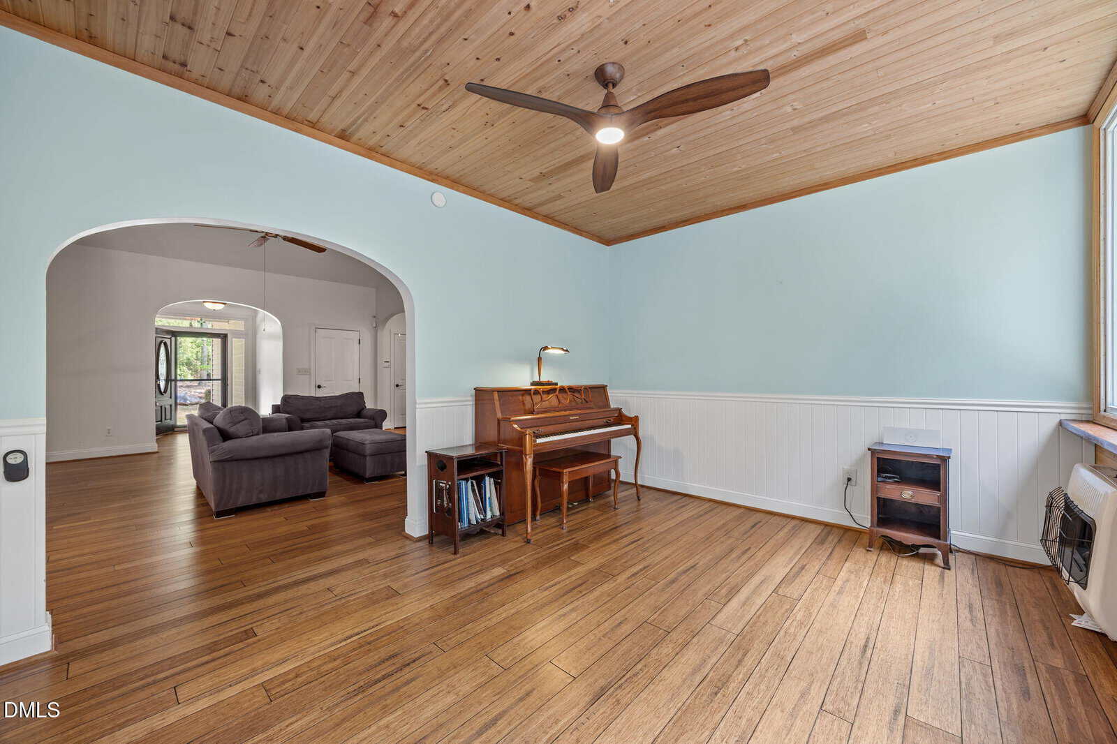 A living room with light blue walls, wood-paneled ceiling, and wood floors. There is a piano with a lamp, a small side table, and a ceiling fan. An open archway leads to a seating area with sofas.