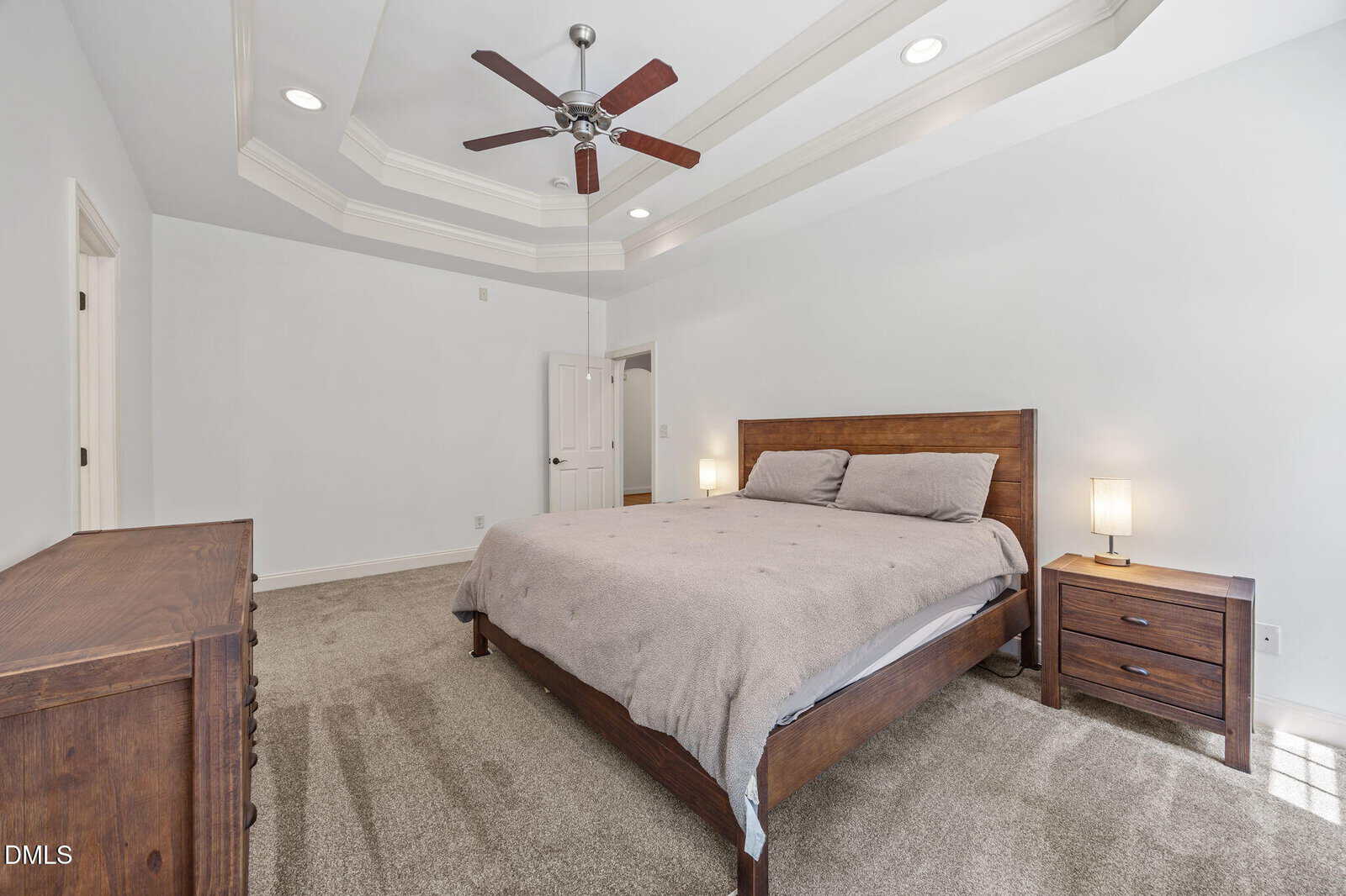 A bedroom with beige carpet, a wooden bed with a beige comforter, two matching nightstands with lamps, a wooden dresser, white walls, a ceiling fan, and a tray ceiling with recessed lighting.