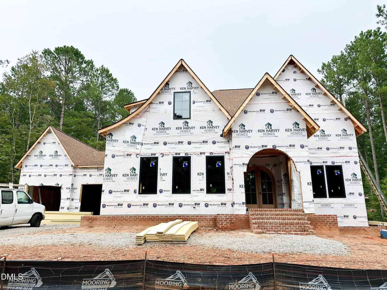 A large two-story house under construction at 300 Ironwood Boulevard, Youngsville, is covered in protective house wrap with visible branding. Bricks, construction materials, and a white van sit in front of the property, which is surrounded by trees and temporary fencing.
