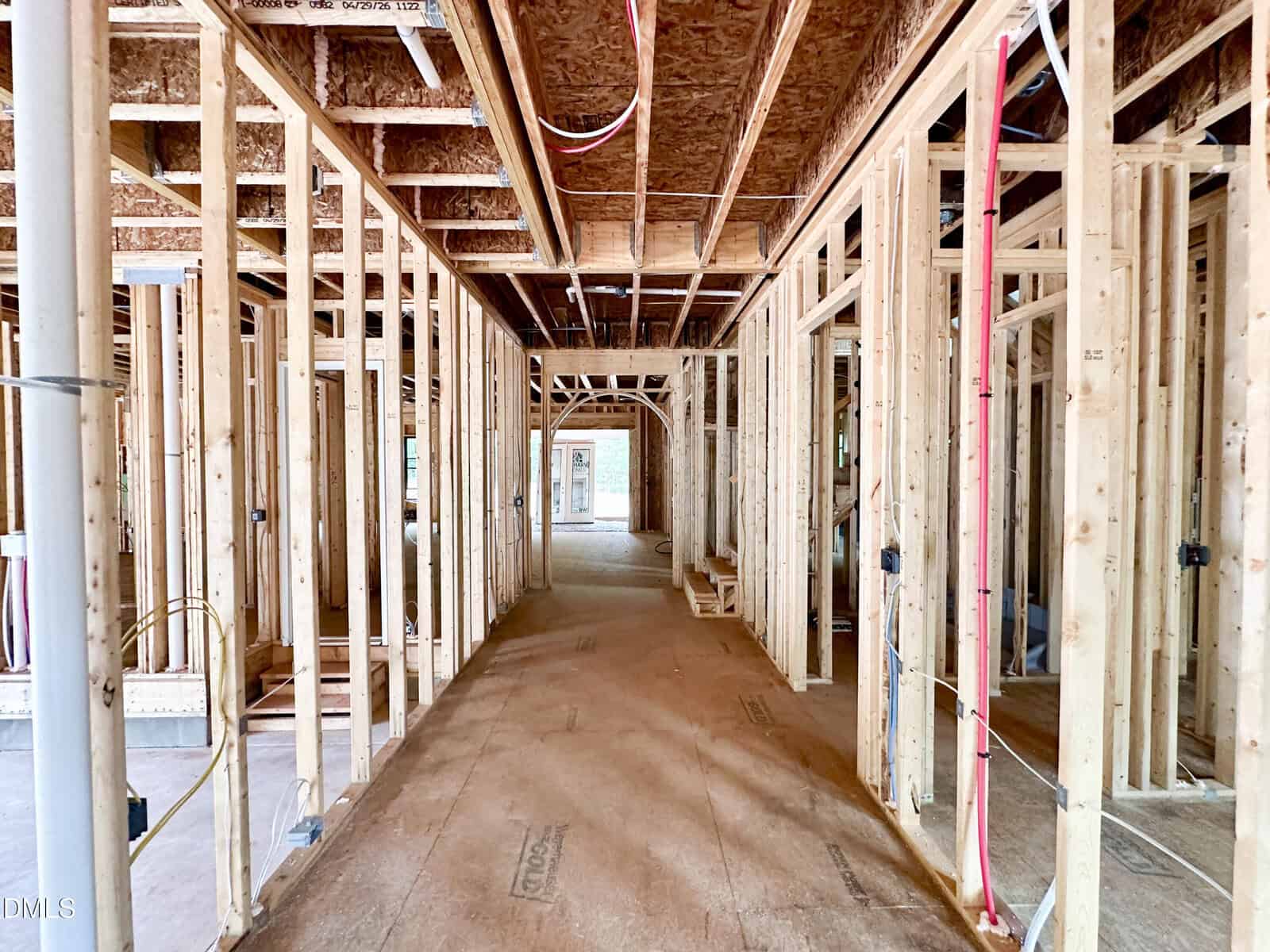 Interior view of a house under construction, showing exposed wooden framing, electrical wiring, plumbing pipes, and unfinished walls and ceiling with natural light coming from windows at the far end.