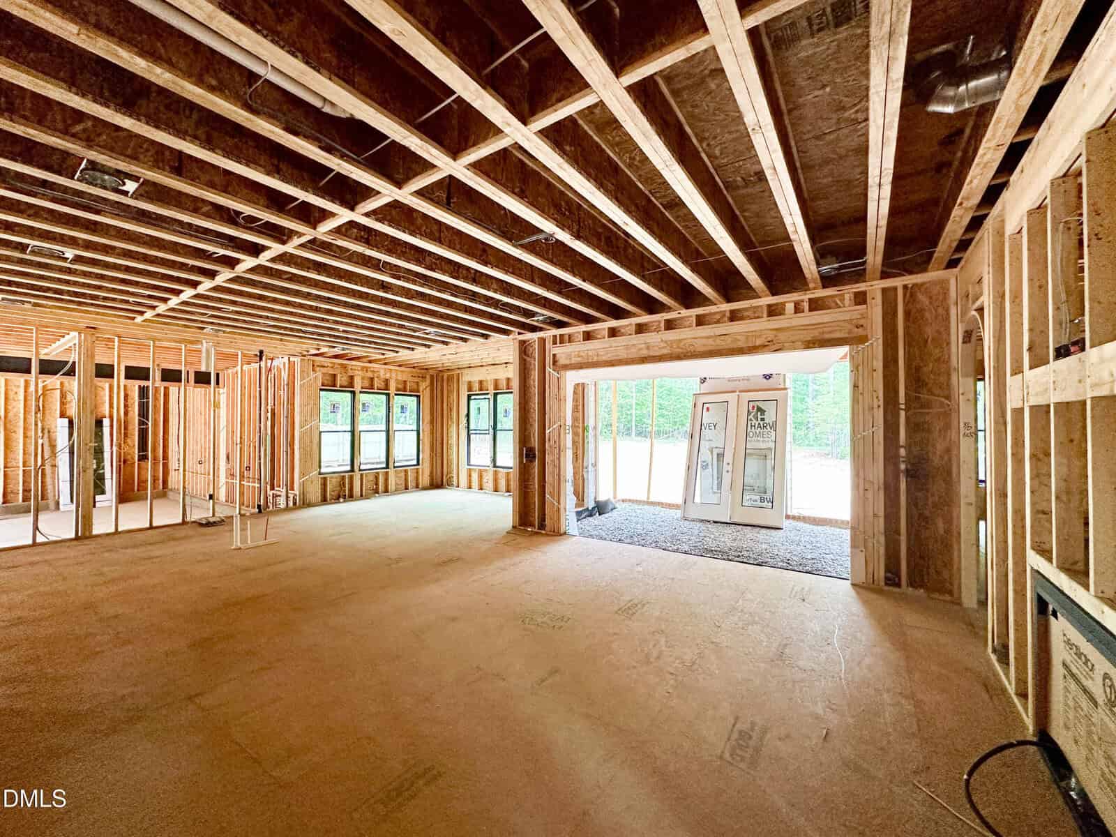 Interior view of a house under construction with exposed wooden framing, unfinished walls, large windows, and an open space leading to a patio door and outside. Natural light fills the empty room.