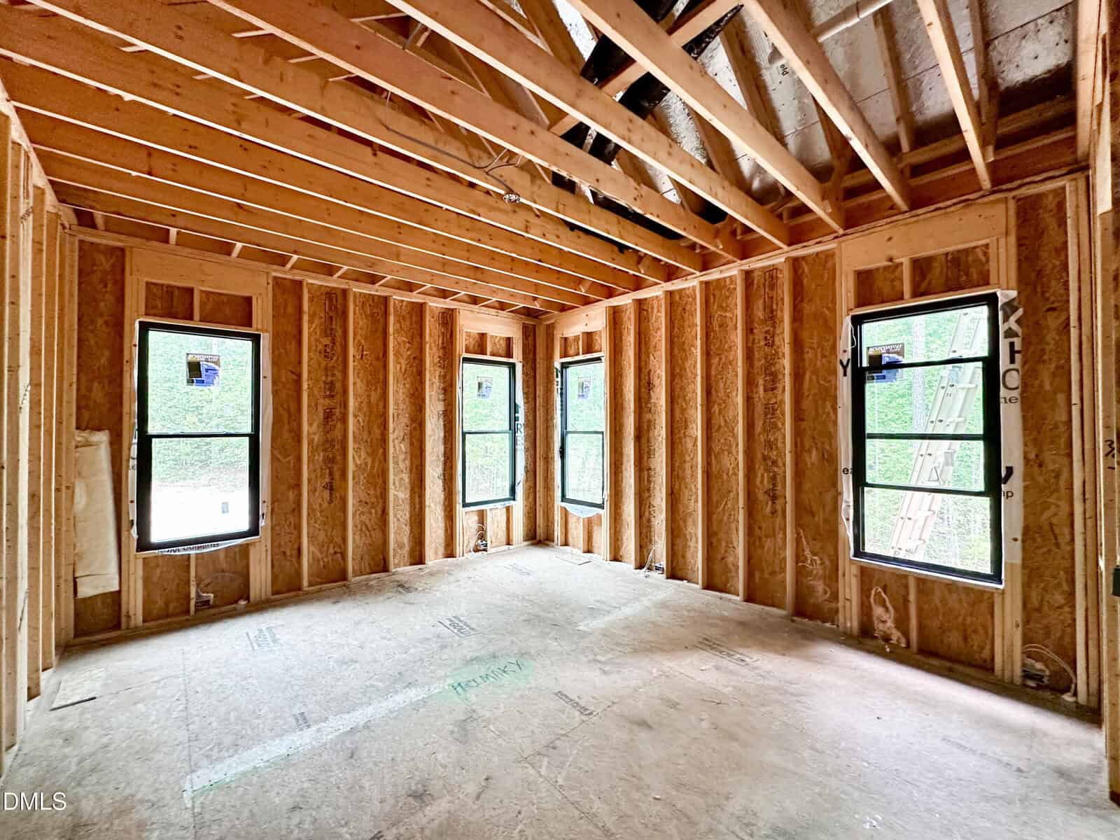 Unfinished room with exposed wooden framing, three windows, and no drywall or flooring; light comes in through the windows, highlighting the beams and open construction.