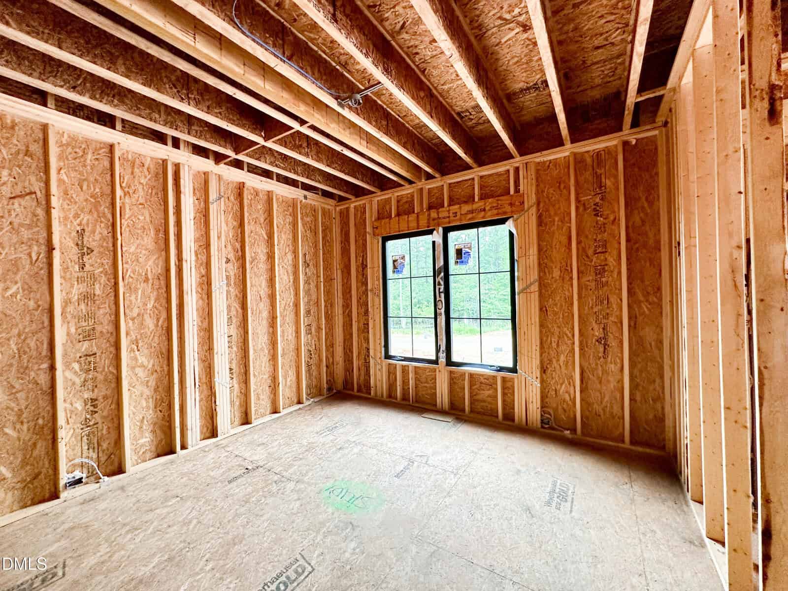 Interior of a house under construction, showing exposed wooden framing, ceiling beams, and an unfinished floor. There are two windows letting in natural light.