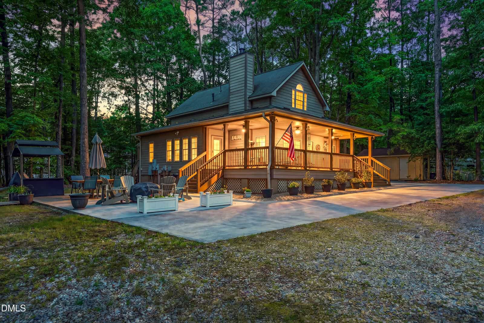 A cozy house at 119 Nashua Drive, Louisburg, glows at dusk with warm interior lights. Surrounded by tall trees, the inviting front porch features a spacious patio with seating, potted plants, and an American flag.
