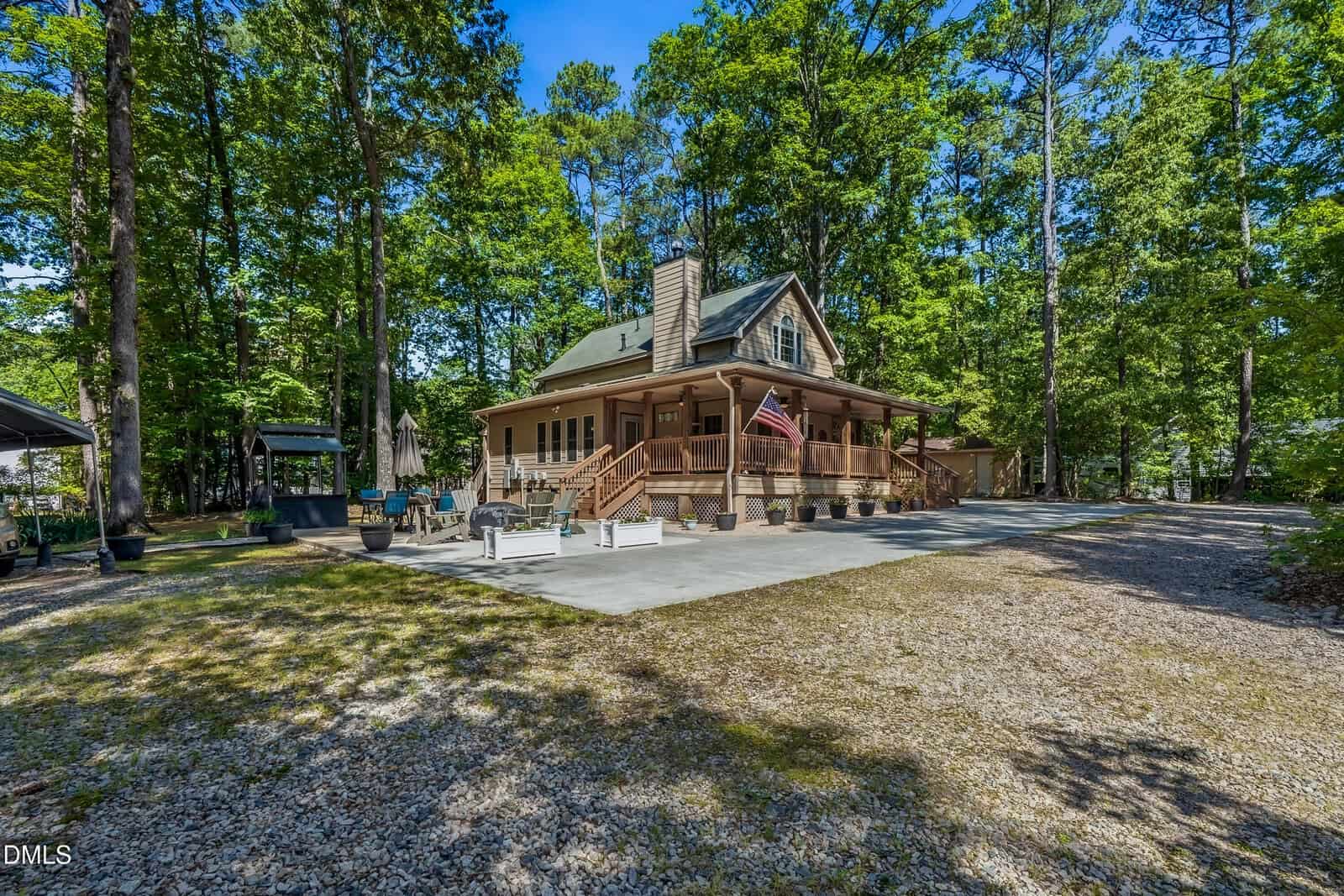 A cozy wooden house with a large covered porch, American flag, and rocking chairs, surrounded by tall trees and a gravel driveway on a sunny day.