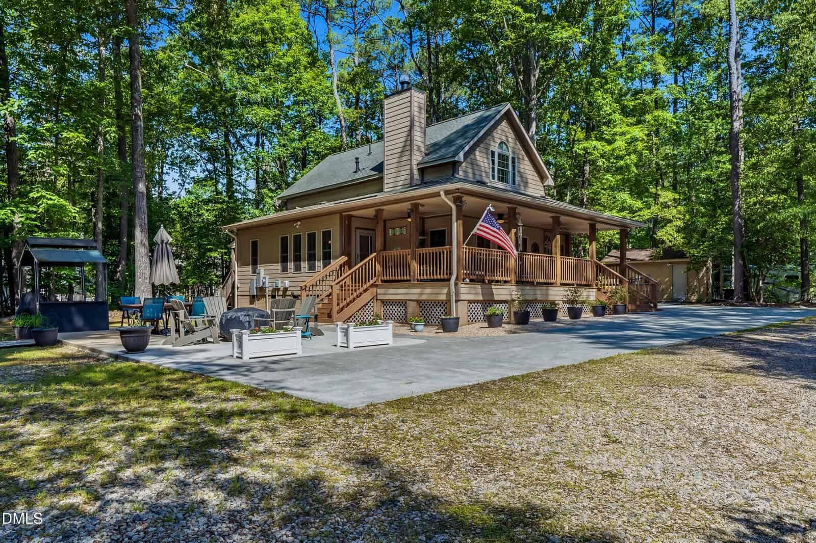 A two-story wooden house with a wraparound porch, American flag, and outdoor seating area, surrounded by trees and greenery on a sunny day.
