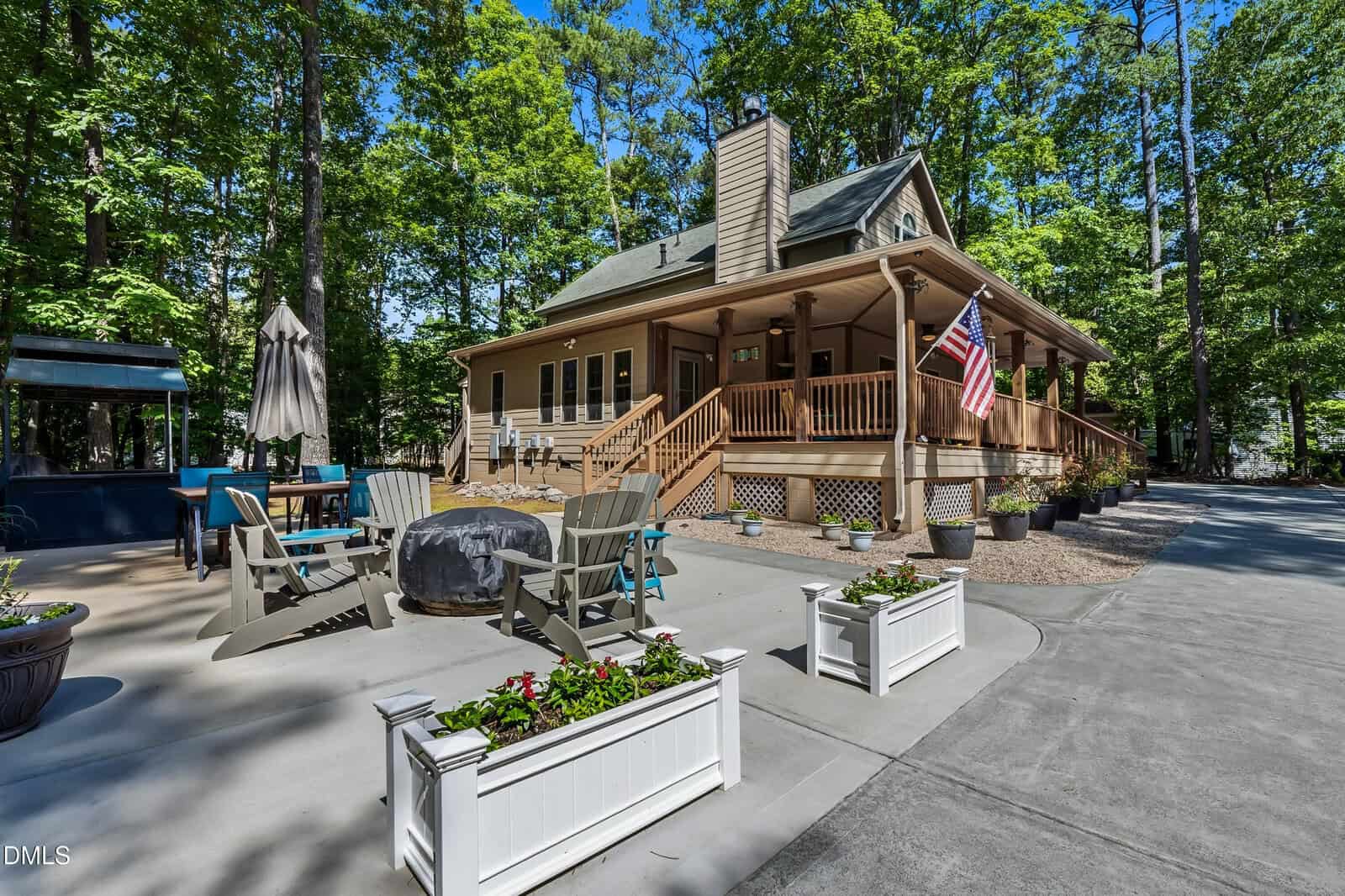 A cozy house with a wraparound porch, American flag, and surrounded by tall trees. The patio has Adirondack chairs around a fire pit, planter boxes with flowers, and outdoor seating under an umbrella.