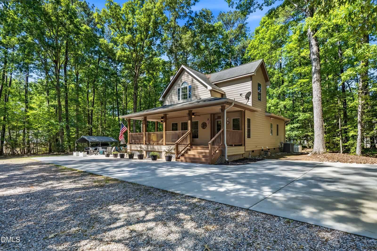 A tan two-story house with a covered front porch and American flag, surrounded by trees. The house has a large driveway and is set on a wooded lot with bright, sunny weather.