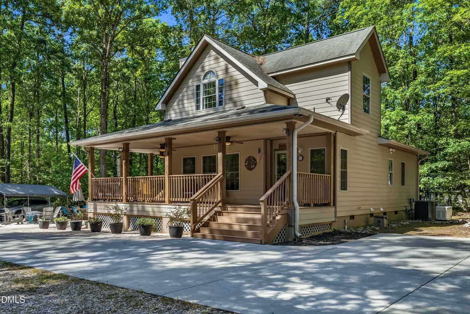 A beige two-story house with a covered front porch, American flag, potted plants, and surrounded by tall green trees. The driveway is paved with concrete, and outdoor furniture is visible in the background.