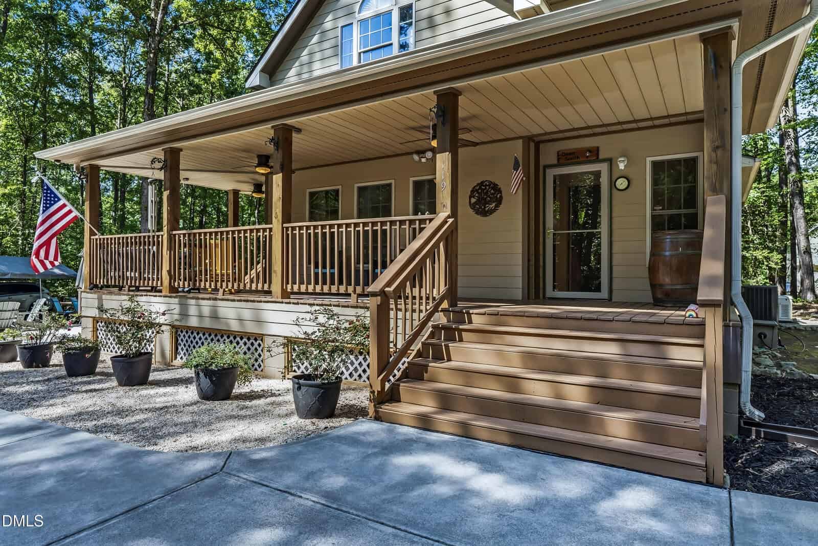 A tan house with a large covered front porch, wooden railings, potted plants, American flags, and stairs, surrounded by trees and bordered by a concrete walkway.