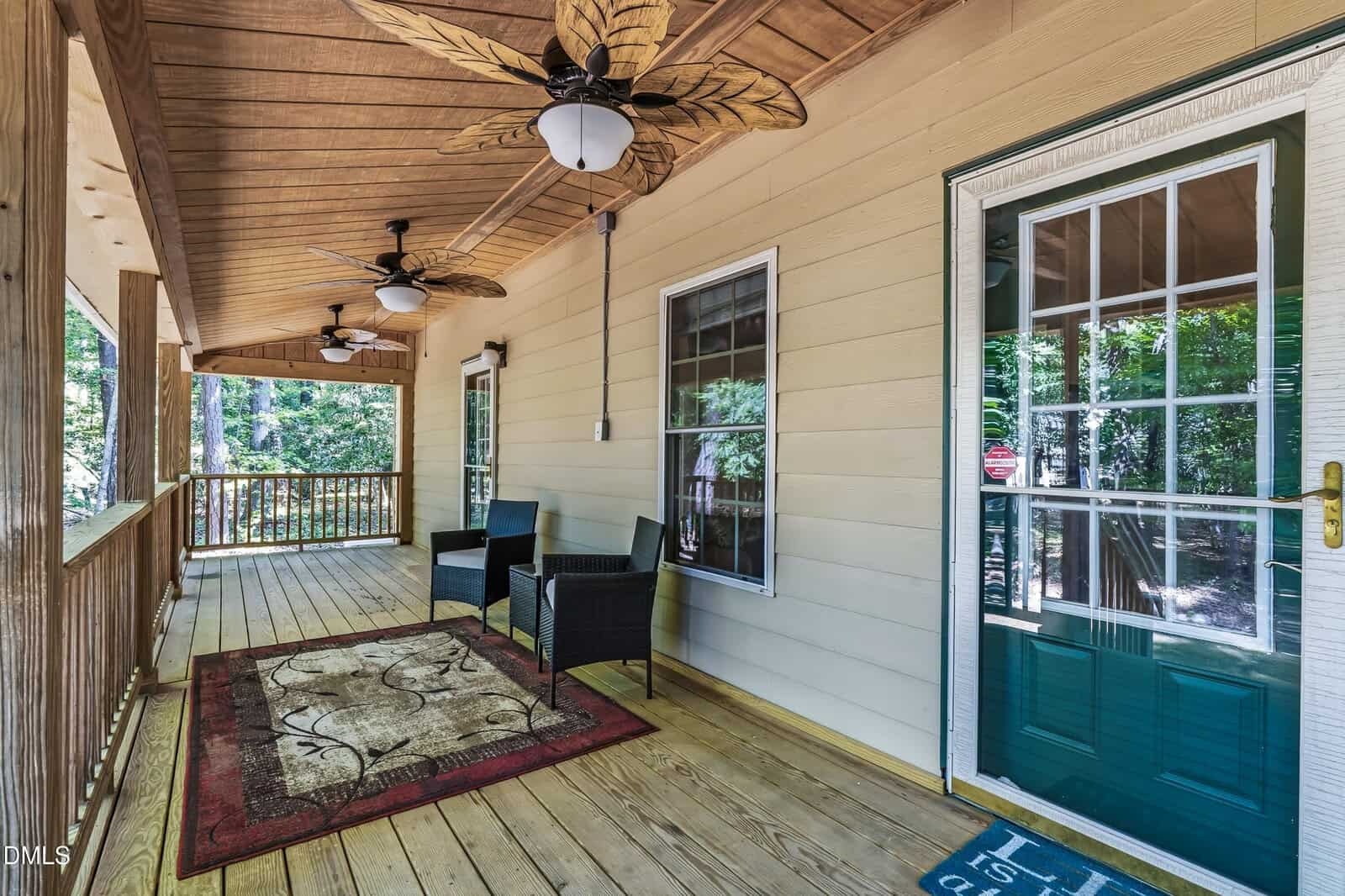 Spacious wooden porch with ceiling fans, two black chairs, and a small table on a patterned rug. Green door and large windows overlook a forested area.