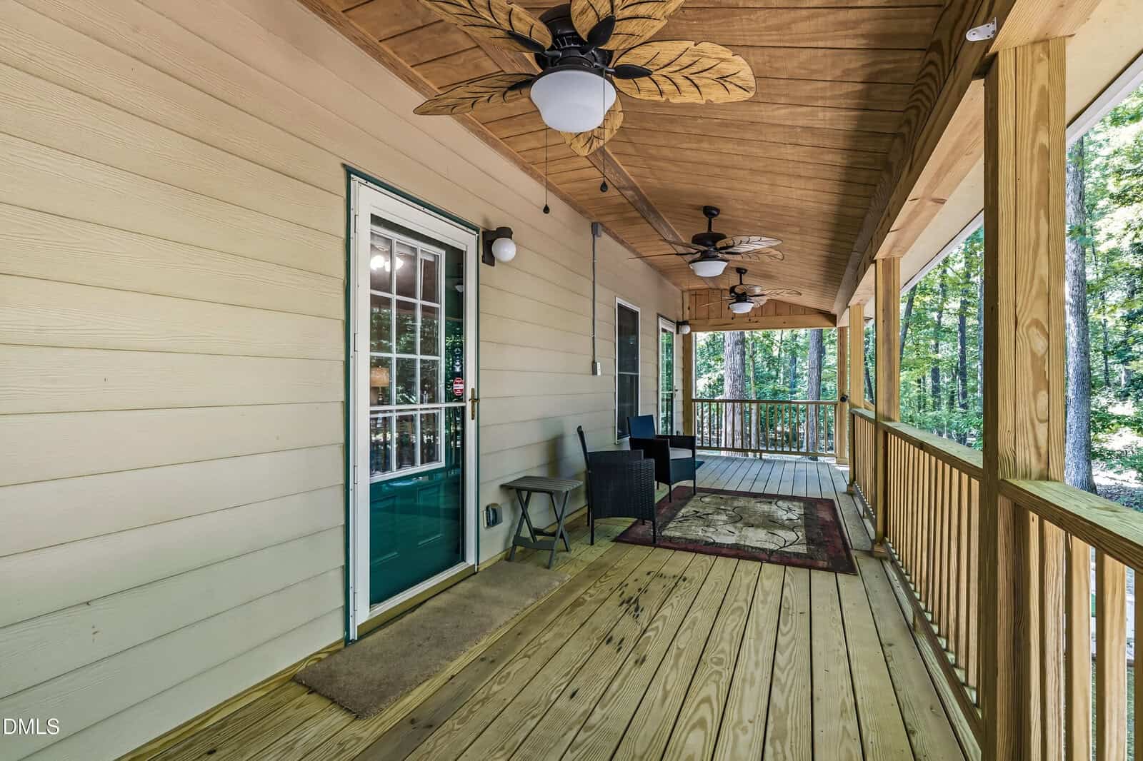 A wooden porch with a ceiling, two ceiling fans, outdoor seating, rug, and railing, attached to a light-colored house and overlooking a wooded area.