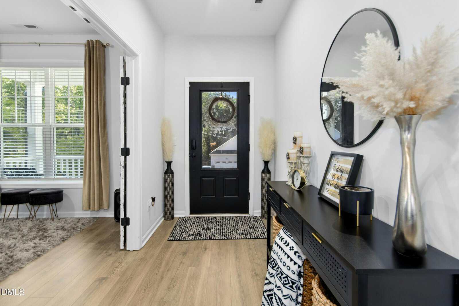 Modern entryway with a black front door, round mirror above a black console table, decorative pampas grass, and baskets beneath the table. A room with large windows and curtains is visible to the left.