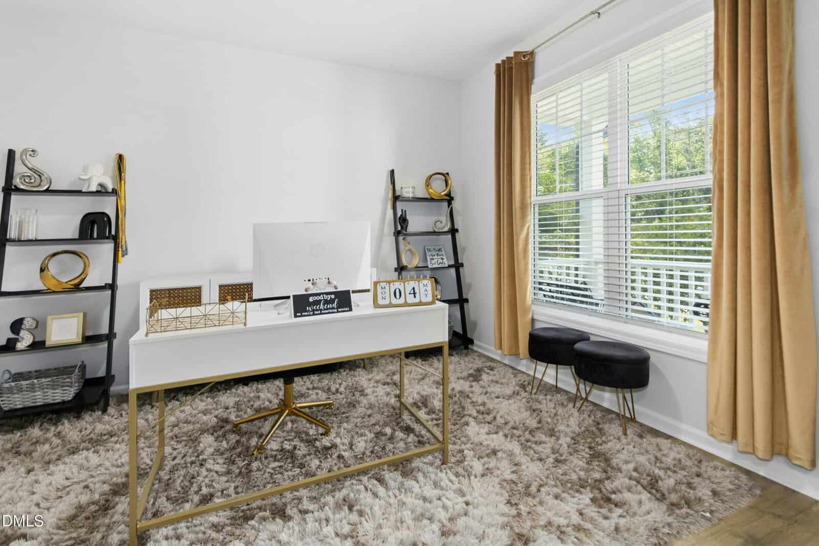 A modern home office with a white desk, a computer, decorative items, black shelving units, gold and black accents, beige curtains, and a large window letting in natural light onto a shaggy rug.