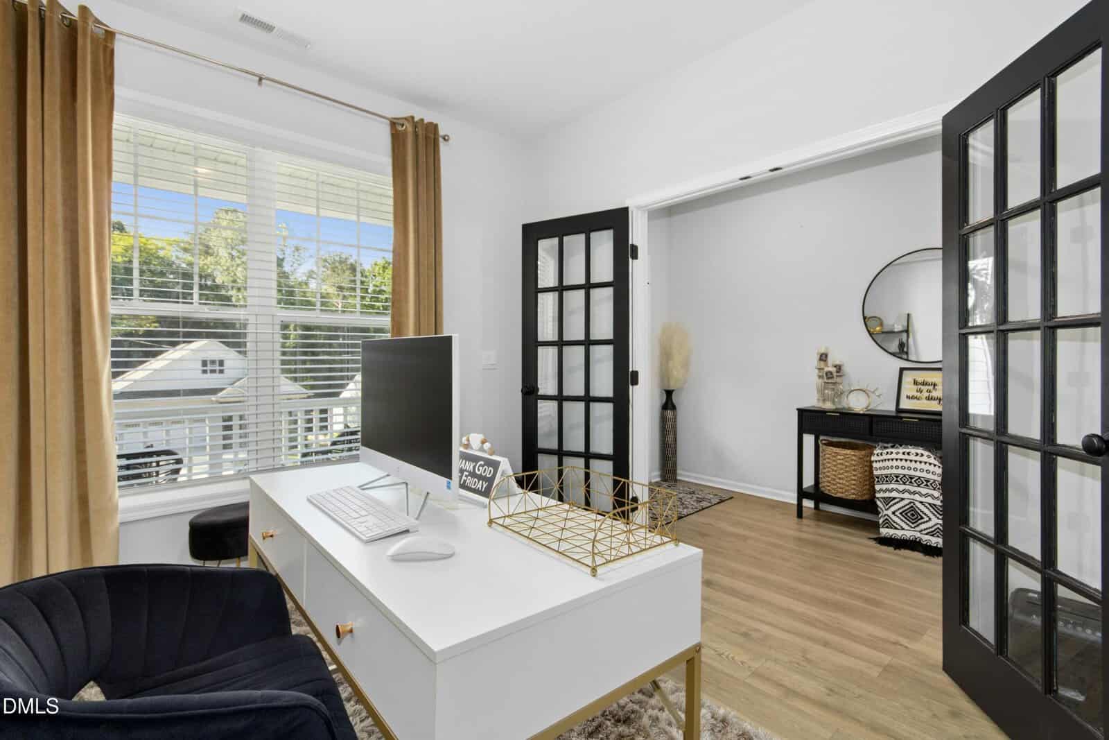 A bright home office with a white desk, computer monitor, and gold accents, featuring black French doors, a large window with brown curtains, and a view of a stylish entryway with decor and a round mirror.