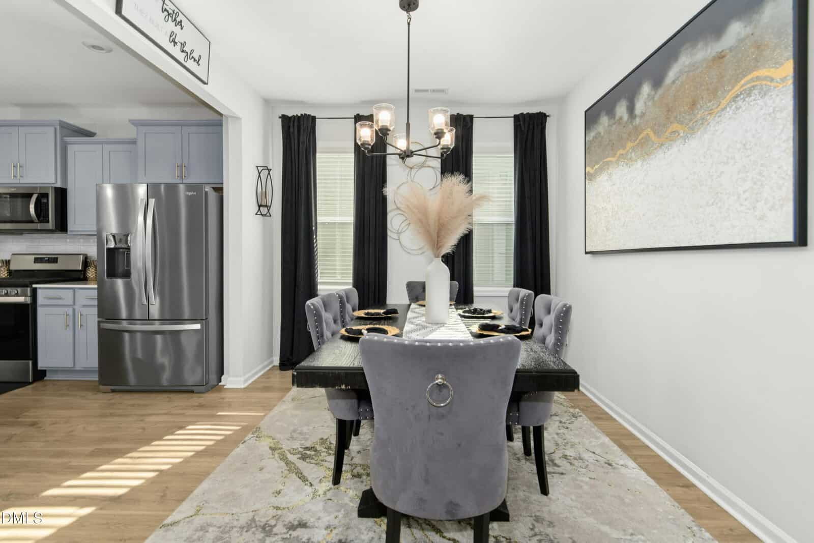 A modern dining room with a black table set for six, gray velvet chairs, a vase with pampas grass centerpiece, large abstract wall art, floor-length black curtains, and a view of a stainless steel kitchen.