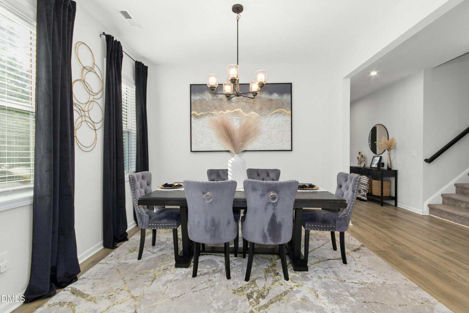 Modern dining room with six gray velvet chairs around a black table, a white vase with pampas grass centerpiece, abstract wall art, a chandelier, and large windows with dark curtains. Stairs and a console table are visible nearby.