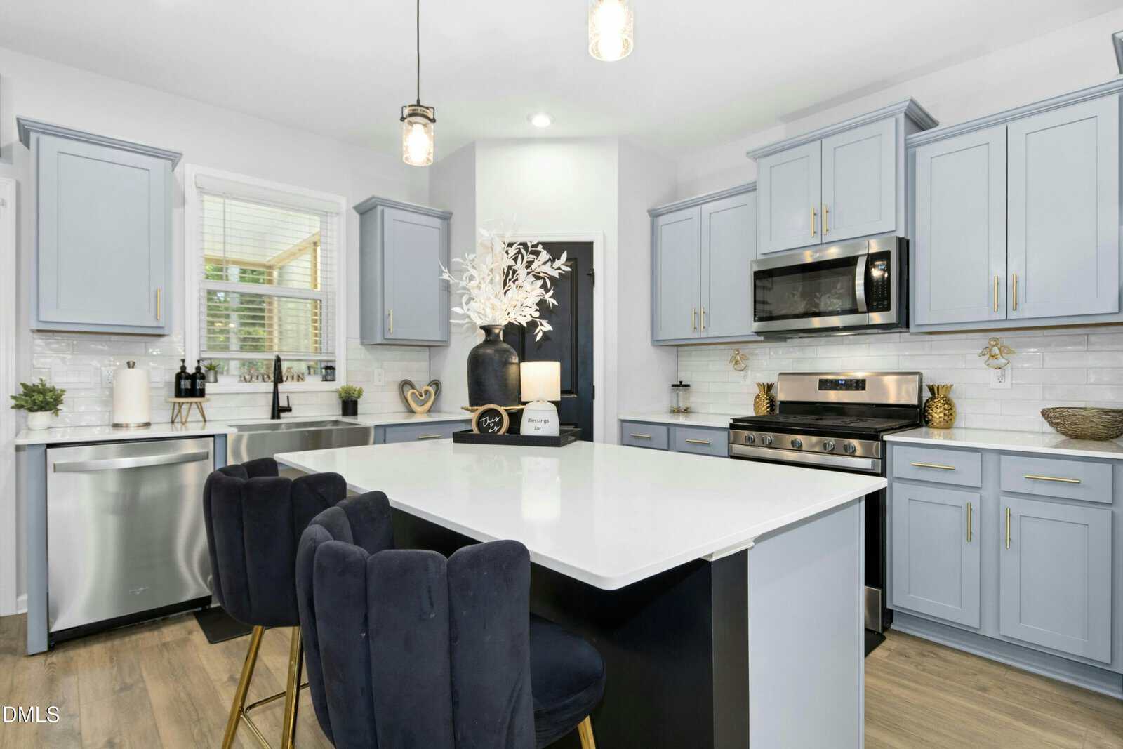 Modern kitchen with light blue cabinets, stainless steel appliances, white countertops, black island with three dark cushioned chairs, decorative vases, and wood flooring. Bright, clean, and stylish atmosphere.