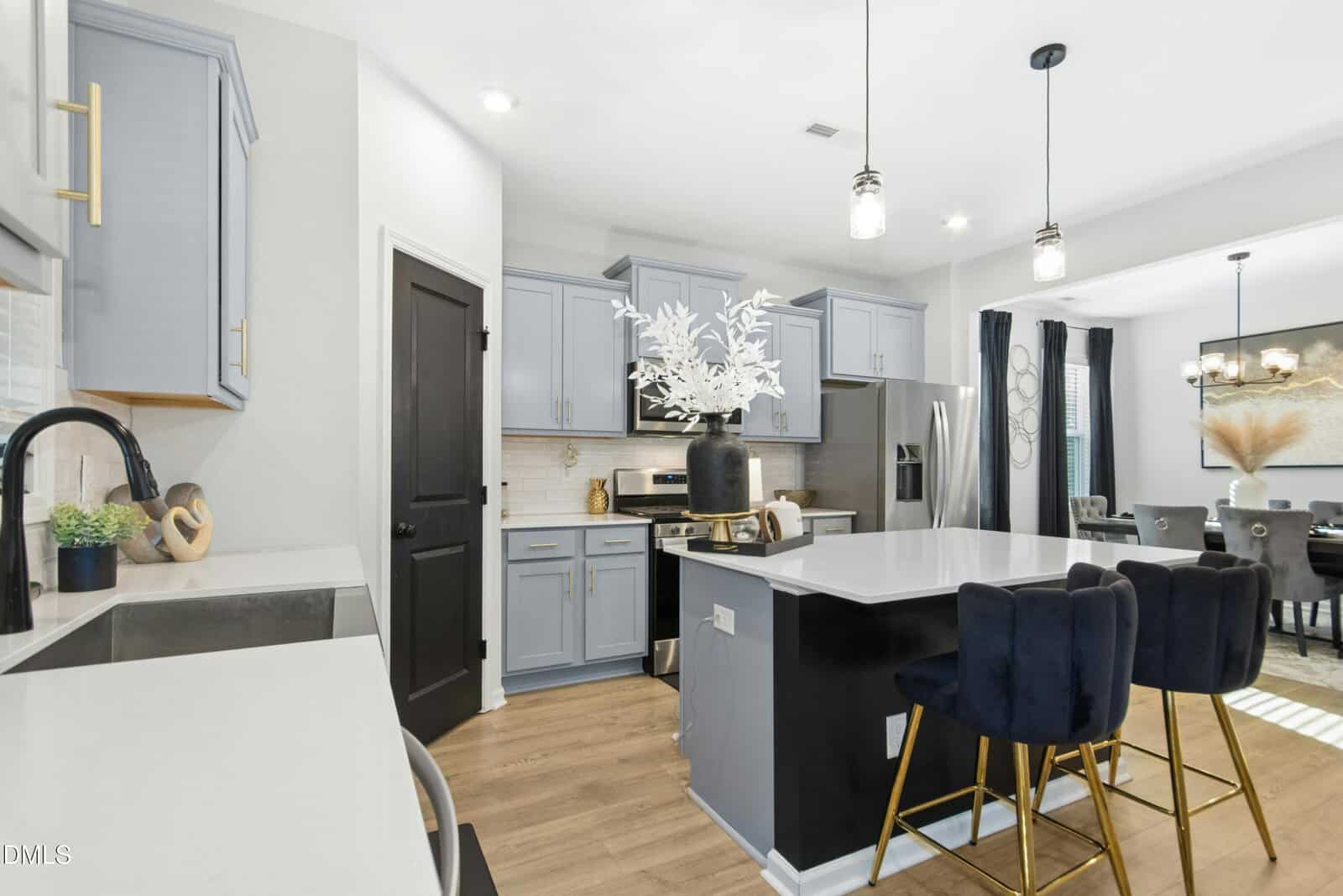 Modern kitchen with light blue cabinets, black accents, island with white countertop, three dark velvet barstools, stainless steel appliances, and open dining area. White decorative vase sits on the island.