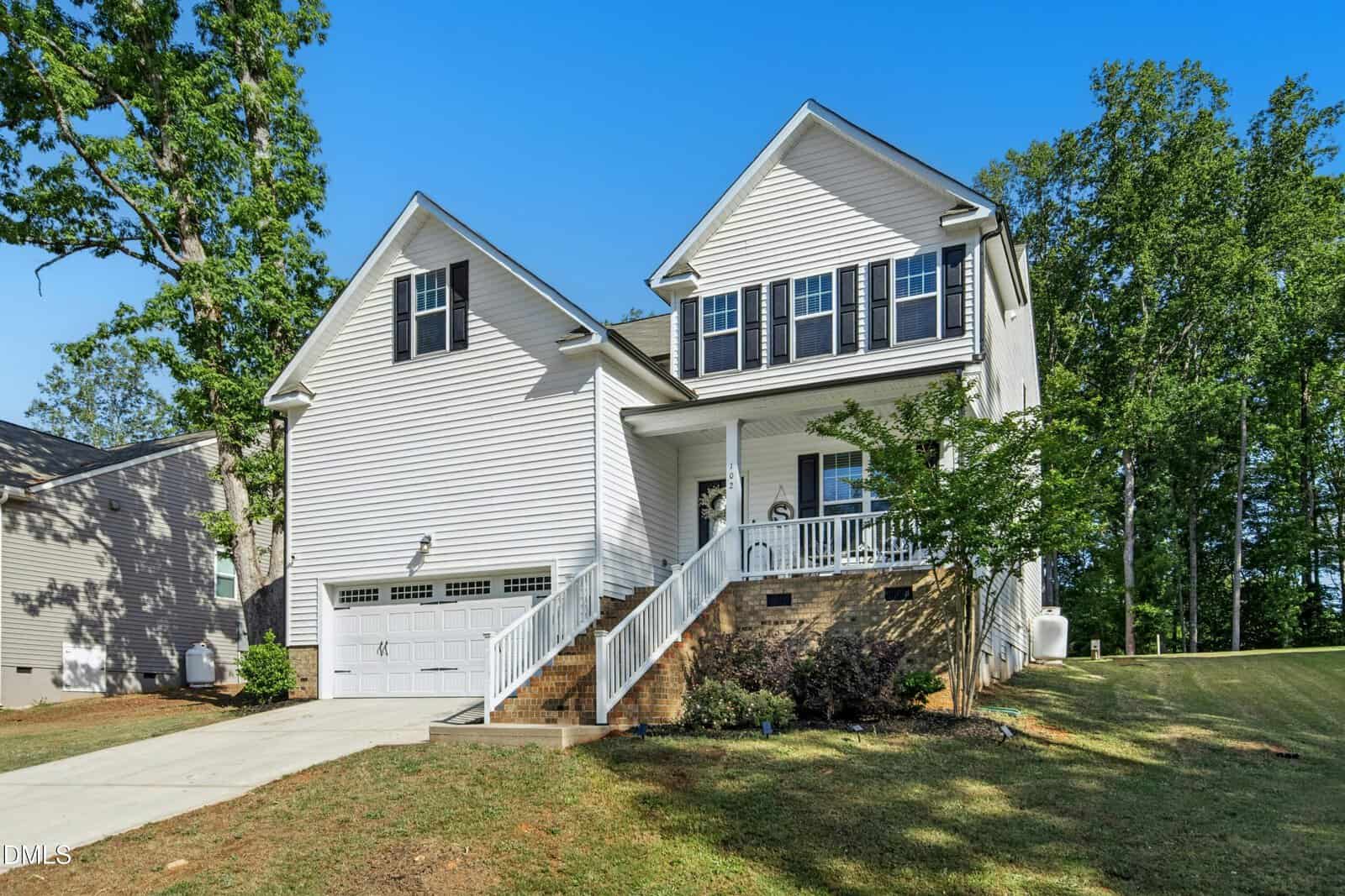 Charming two-story white house with black shutters at 102 Red Dog Drive, Louisburg, featuring an attached two-car garage, a front porch with chairs, brick steps, sloped driveway, and a grassy yard surrounded by trees.