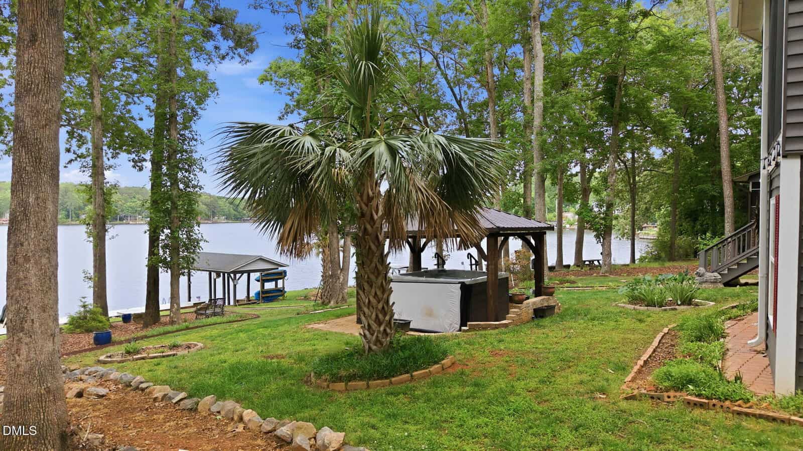 A lakeside backyard with a palm tree, hot tub, and covered seating areas among green grass and trees, overlooking a calm lake. A boat dock is visible in the background under a blue sky.