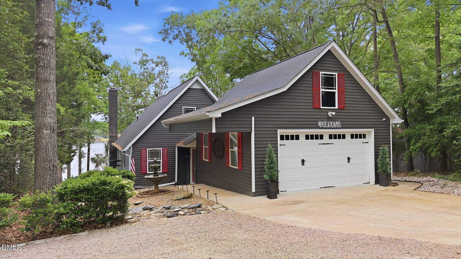 A gray two-story house with white trim and red shutters at 653 Sagamore Drive, Louisburg. Features a single-car garage, sloped driveway, mature landscaping, and a glimpse of the lake in the background.