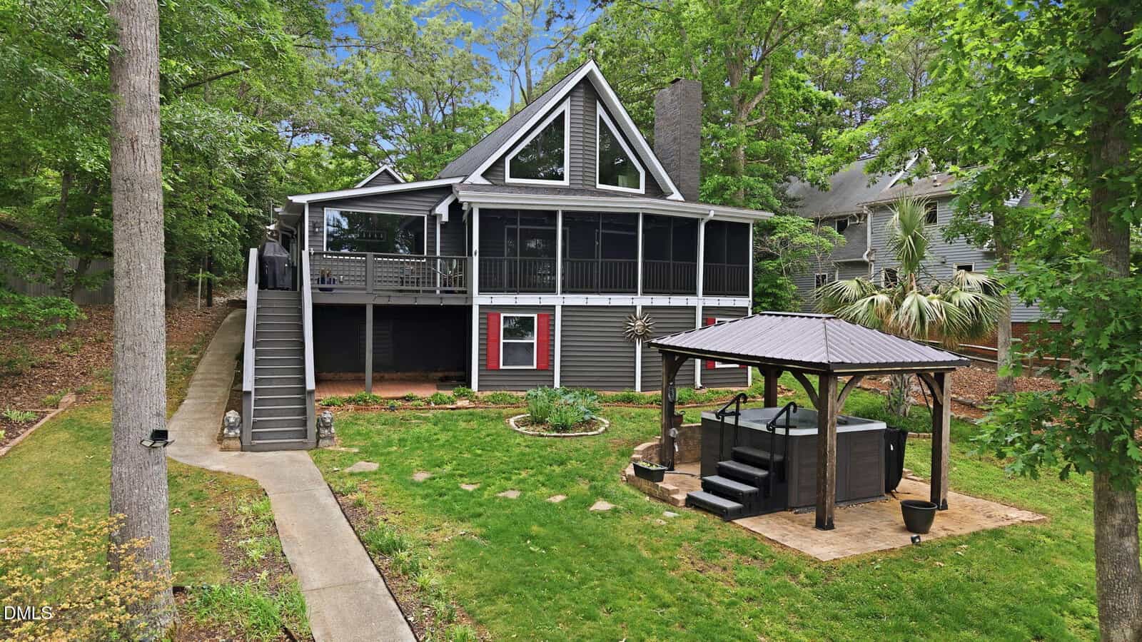 A two-story gray house with a large screened porch, surrounded by trees, features an outdoor staircase, a lawn, and a gazebo with a hot tub on a stone patio in the backyard.