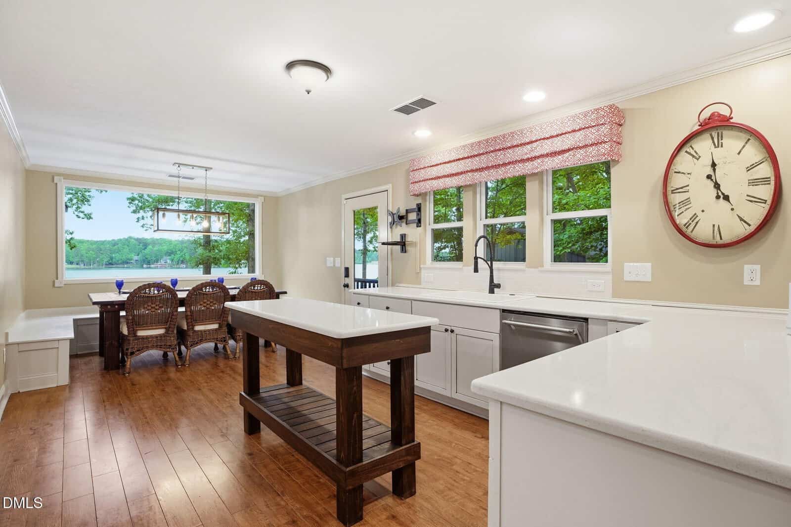 Bright kitchen with wood floors, white cabinets, a center island, farmhouse sink, large wall clock, and windows overlooking trees; dining area with a wooden table and four chairs near a built-in bench is visible.