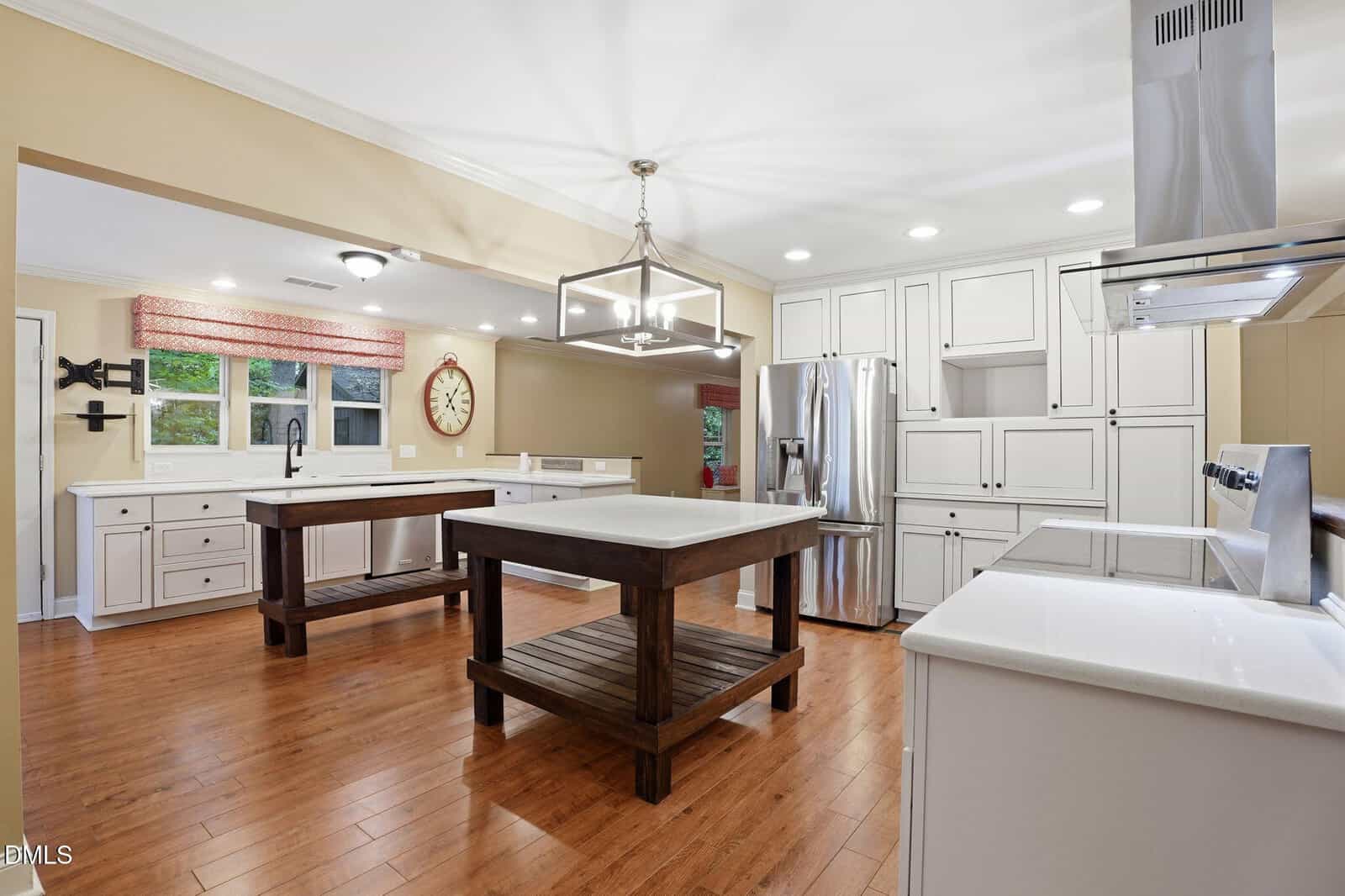 Spacious kitchen with wood floors, white cabinets, stainless steel appliances, two wooden islands, a farmhouse sink under a window, wall clock, and recessed lighting.