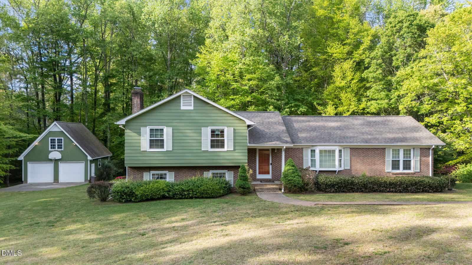 A split-level house with green siding and brick facade, white trim, and a shingled roof sits in front of a wooded area. A detached two-car garage with a basketball hoop is on the left. The lawn is spacious and well-kept.