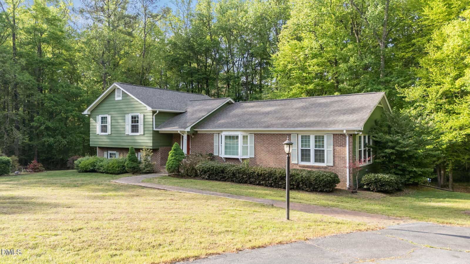 A split-level house with green siding and red brick exterior, surrounded by bushes and trees, sits on a grassy lawn with a curved driveway and a black lamp post in front.