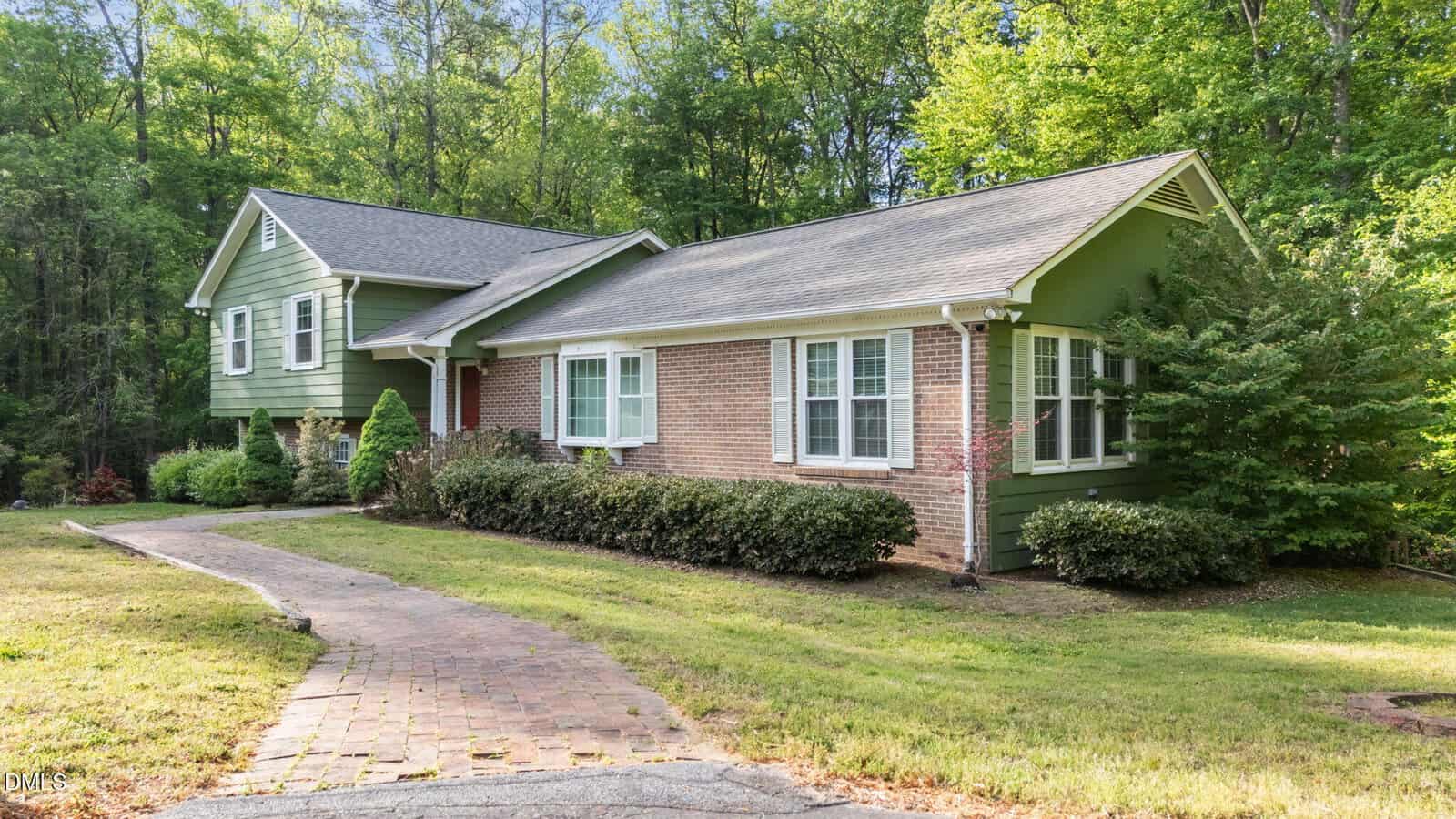 A split-level house with green siding and brick exterior, surrounded by bushes and trees, with a curved brick walkway leading to the front entrance and a grassy lawn in front.