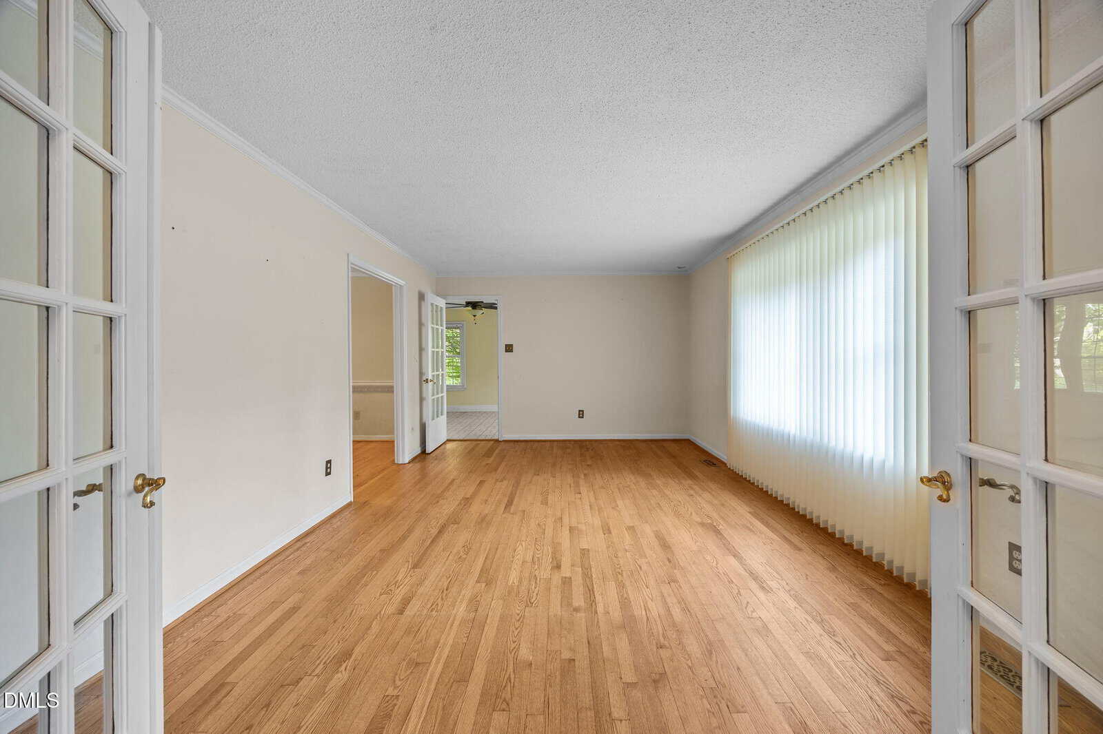 Bright, empty room with hardwood floors, white walls, French doors, and large window covered by vertical blinds, letting in natural light.