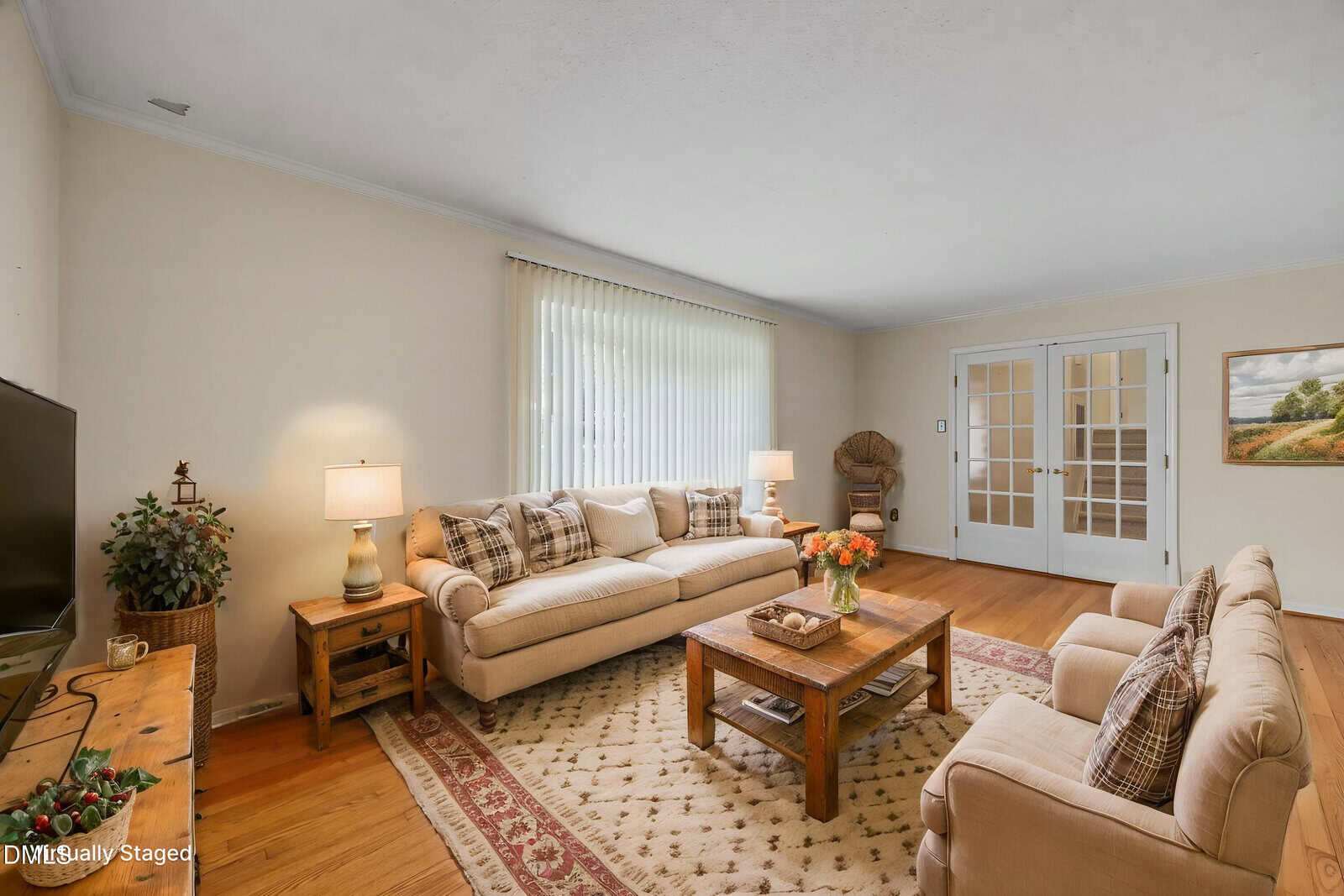 Bright living room with beige walls, two sofas, a wooden coffee table, patterned rug, side tables with lamps, a TV, French doors, and a window with vertical blinds. A framed landscape painting hangs on the right wall.