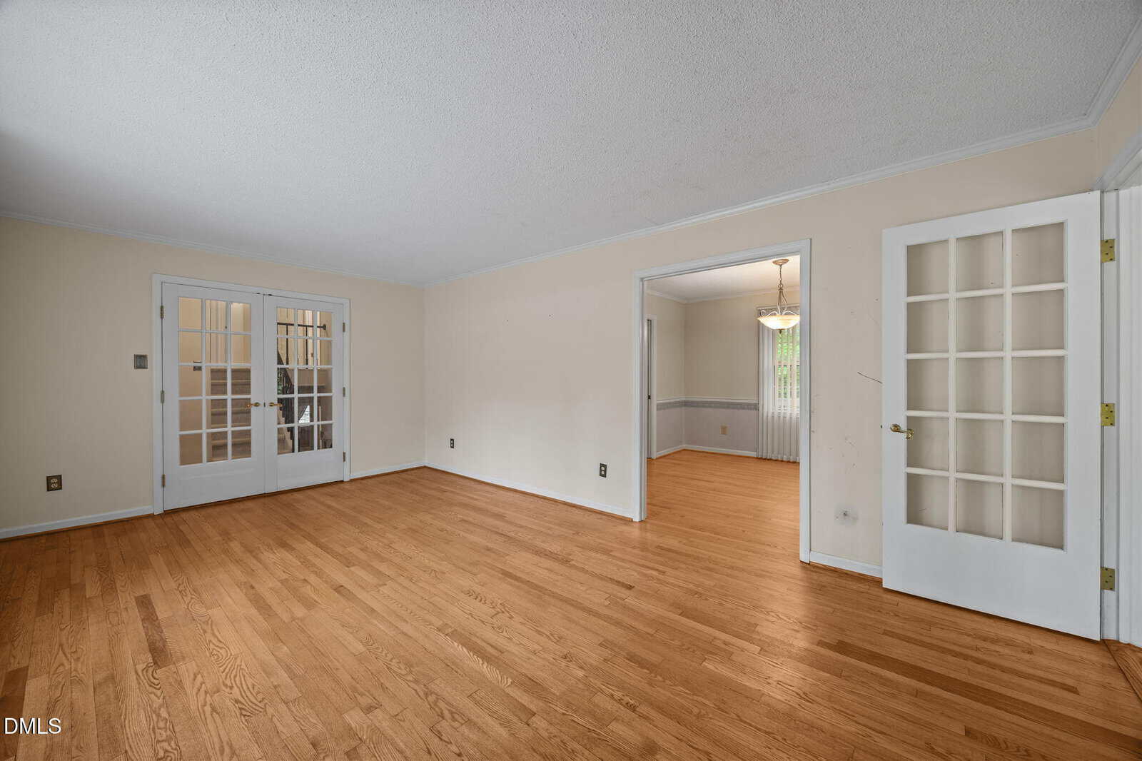 Spacious empty room with light wood flooring, beige walls, white trim, and French doors leading to another room and a dining area with a chandelier.