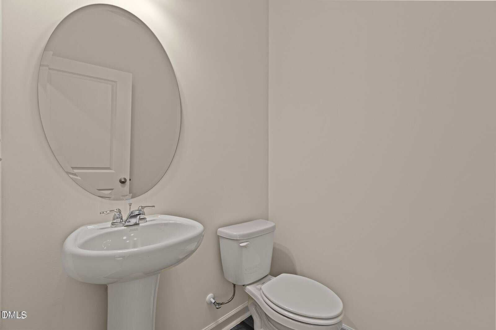 A minimalist bathroom with a pedestal sink, an oval mirror above it, a toilet, and neutral-colored walls. The door is reflected in the mirror.