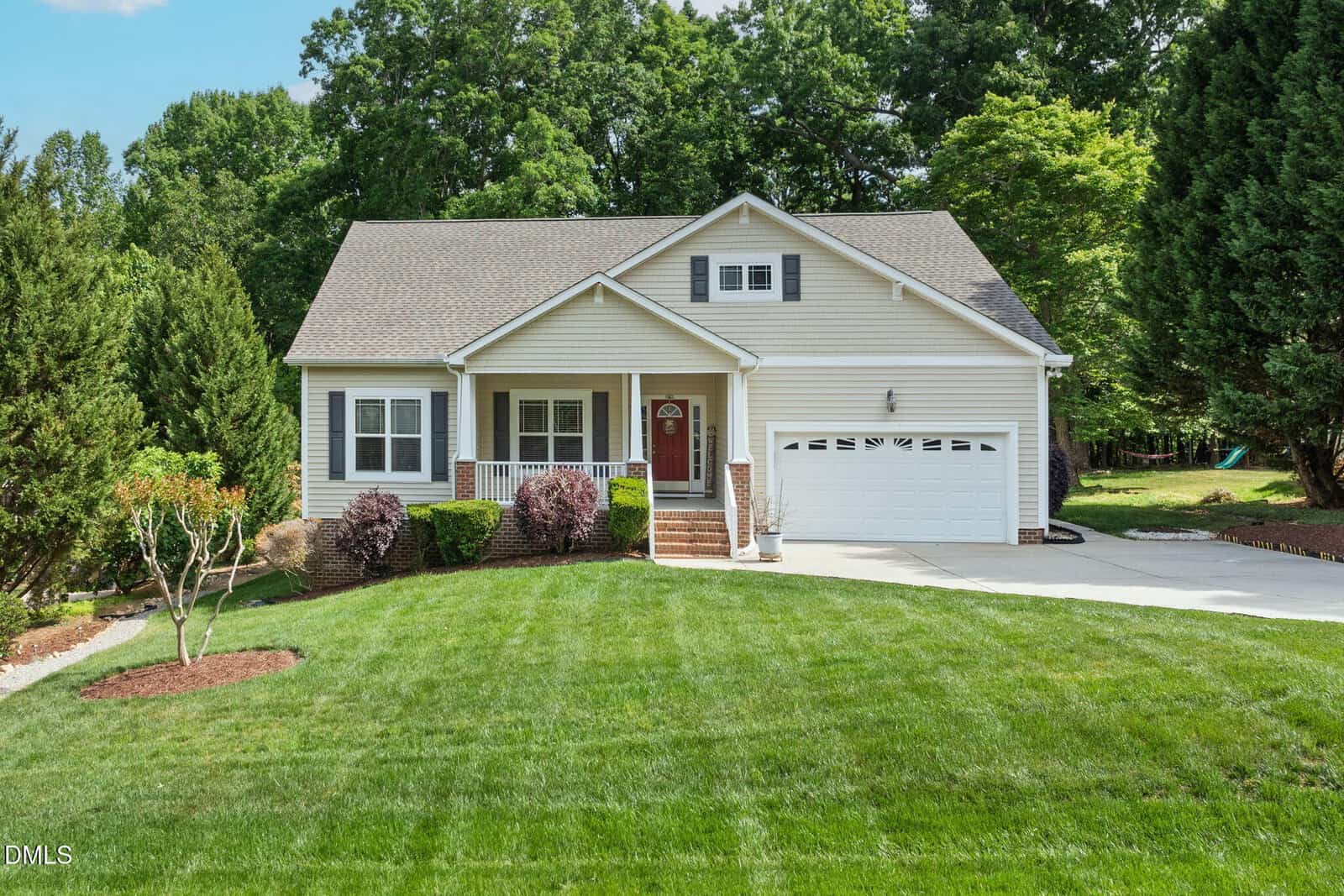 Single-story house with light siding, a covered front porch, and a two-car garage. The home is surrounded by well-maintained landscaping and green lawn, with trees in the background and a concrete driveway in front.