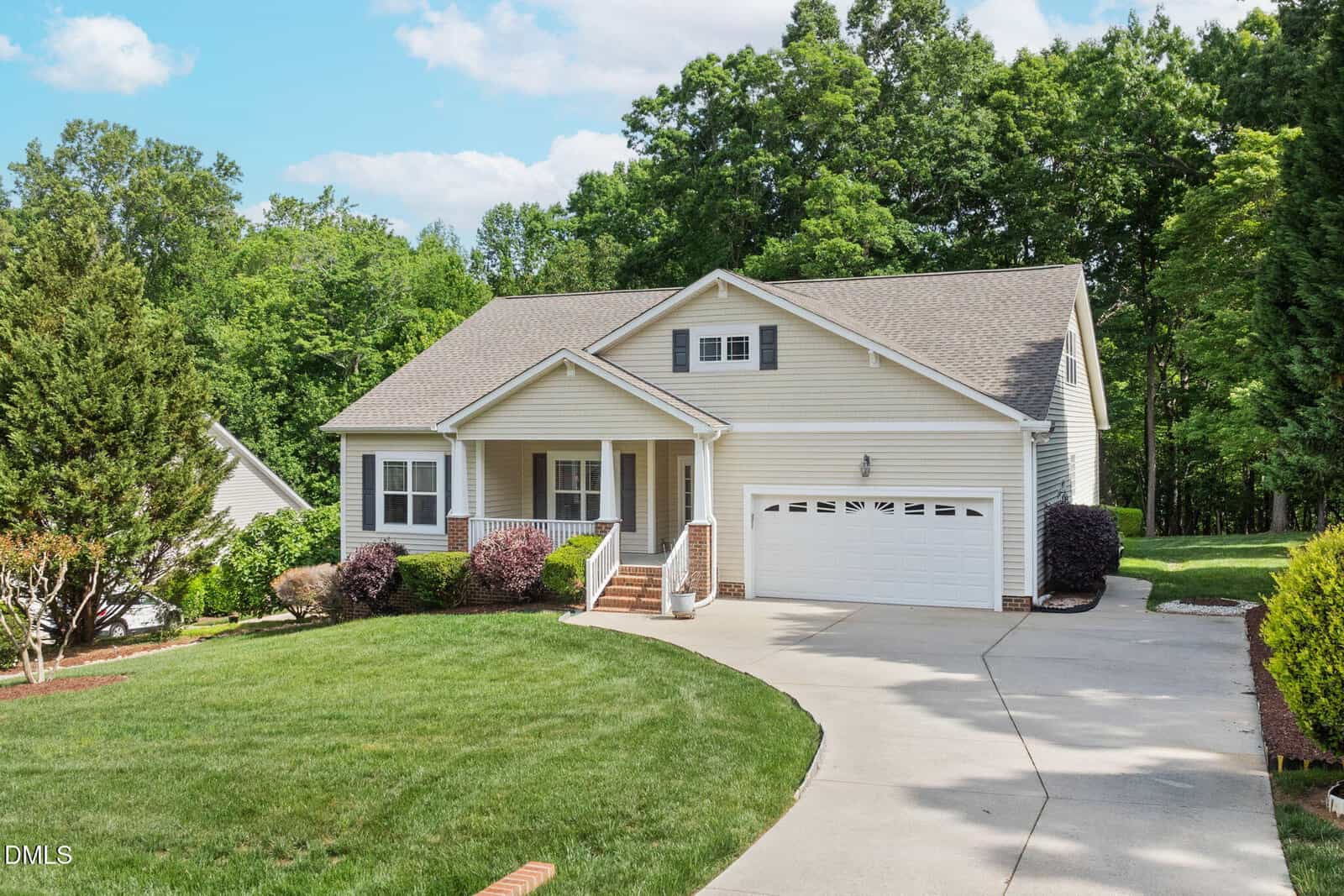 Single-story beige house with a covered front porch, white trim, and attached two-car garage. Curved driveway and well-maintained lawn with shrubs and trees surround the home. Tall trees line the background.