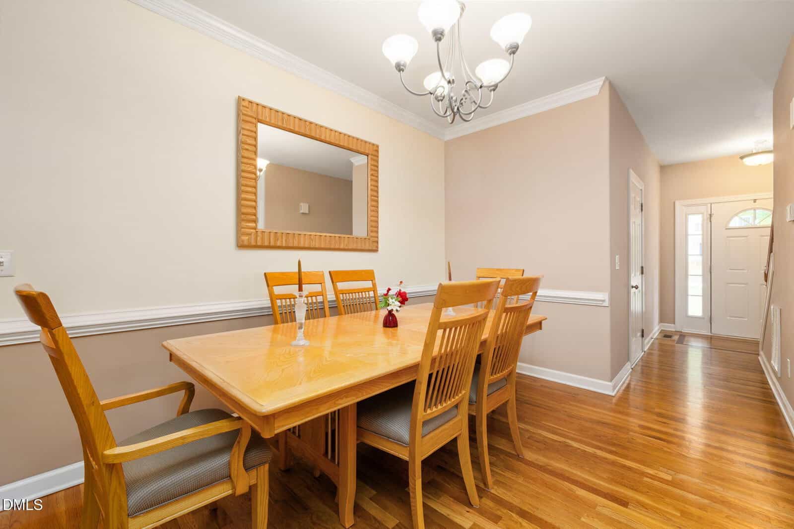 A dining room with a wooden table and six matching chairs, a mirror on the beige wall, hardwood floors, and a chandelier overhead. The space leads to a hallway with the front door visible in the background.