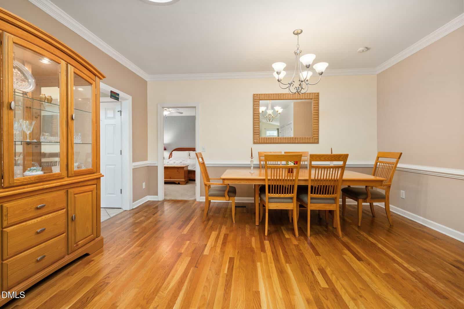 A dining room with a wooden table and six chairs, a chandelier overhead, a large mirror on the wall, and a glass-fronted china cabinet on the left; a bedroom is visible through an open doorway.