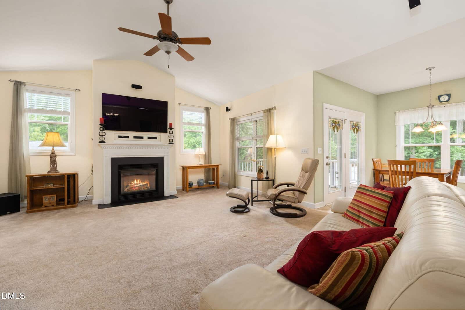 A cozy living room with a beige carpet, white sofa with colorful pillows, fireplace under a wall-mounted TV, recliner chairs, and large windows. Dining area with wooden table and chairs visible in the background.