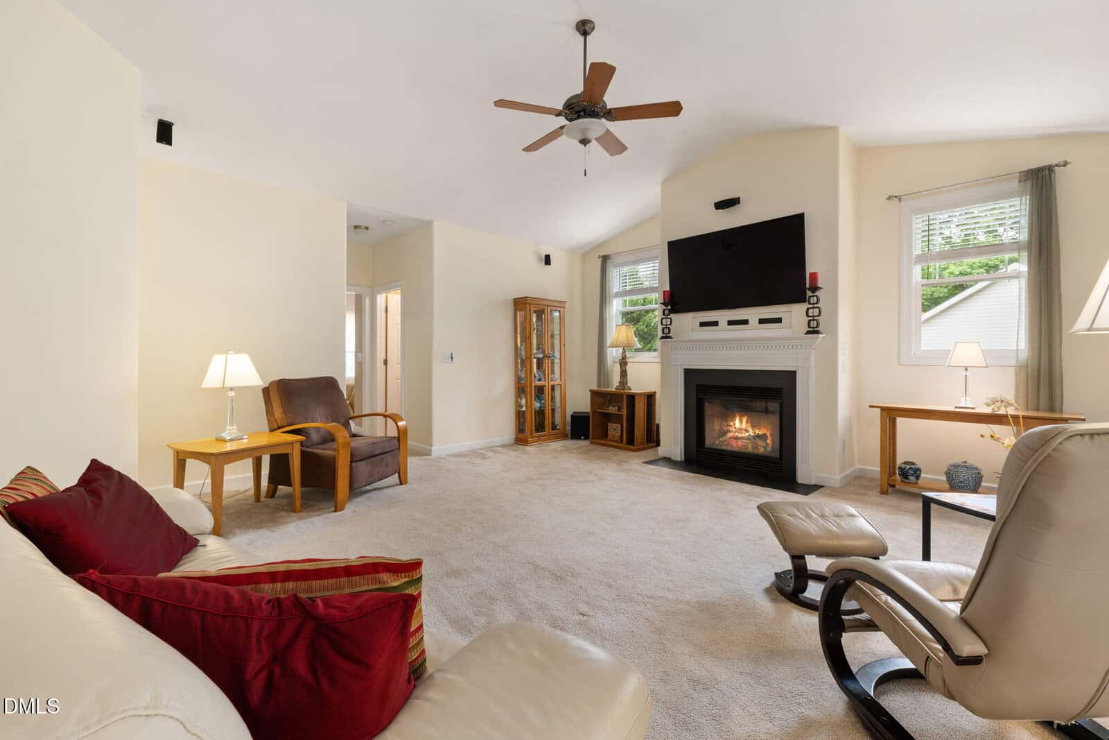 Bright living room with beige carpet, cream walls, a fireplace below a mounted TV, ceiling fan, leather seating, side tables with lamps, and windows with natural light.