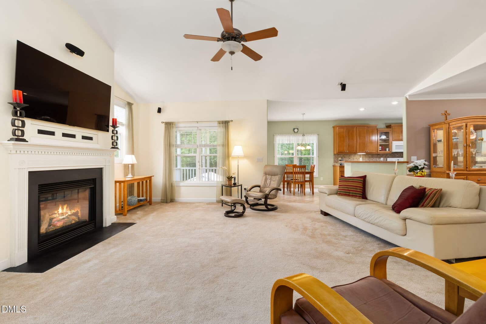 A cozy living room with a fireplace, TV, beige sofas, and an armchair. Large windows let in light. The room opens to a dining area and kitchen with wooden cabinets and a chandelier.