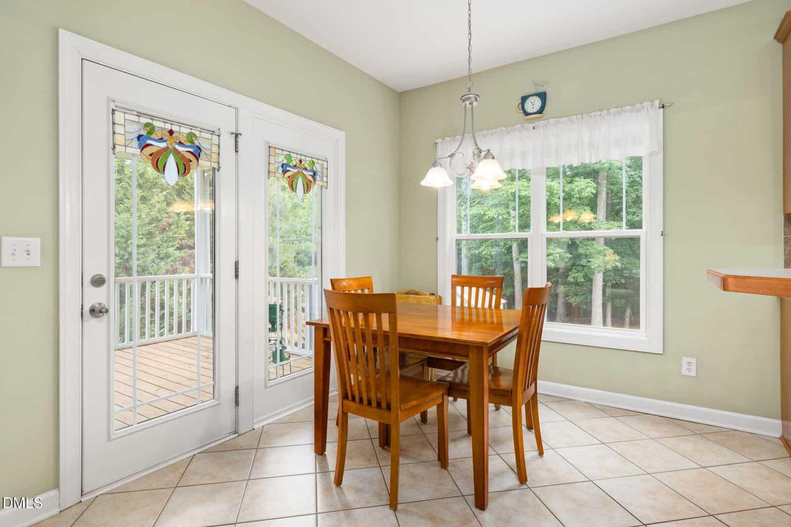 A wooden dining table with four chairs sits by a window and glass doors with stained glass accents, opening to a deck. The room has tiled floors and light green walls.