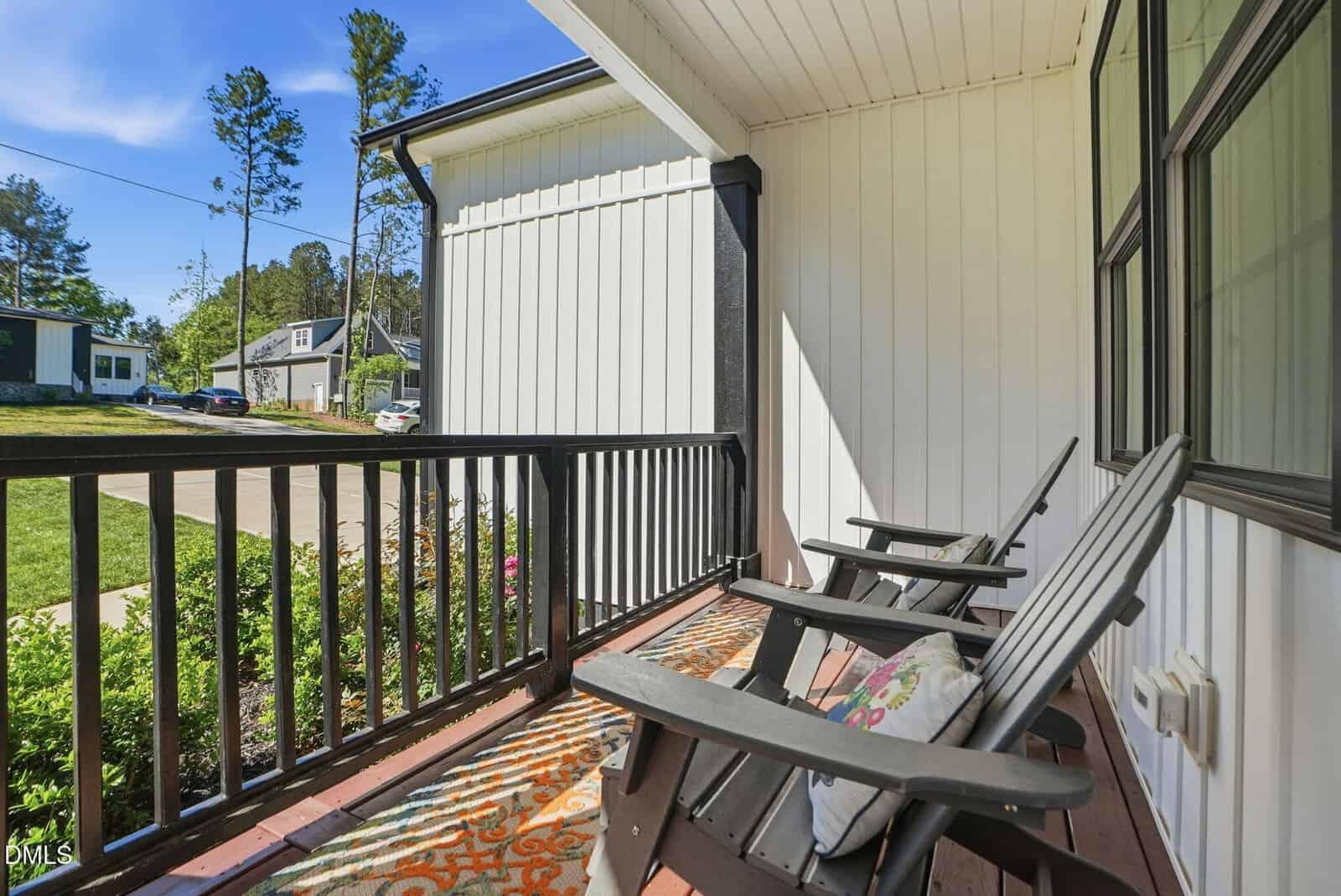 A sunny front porch with two black rocking chairs, a patterned outdoor rug, and pillows. The porch has white siding and black railings, overlooking a green yard and a quiet suburban street.