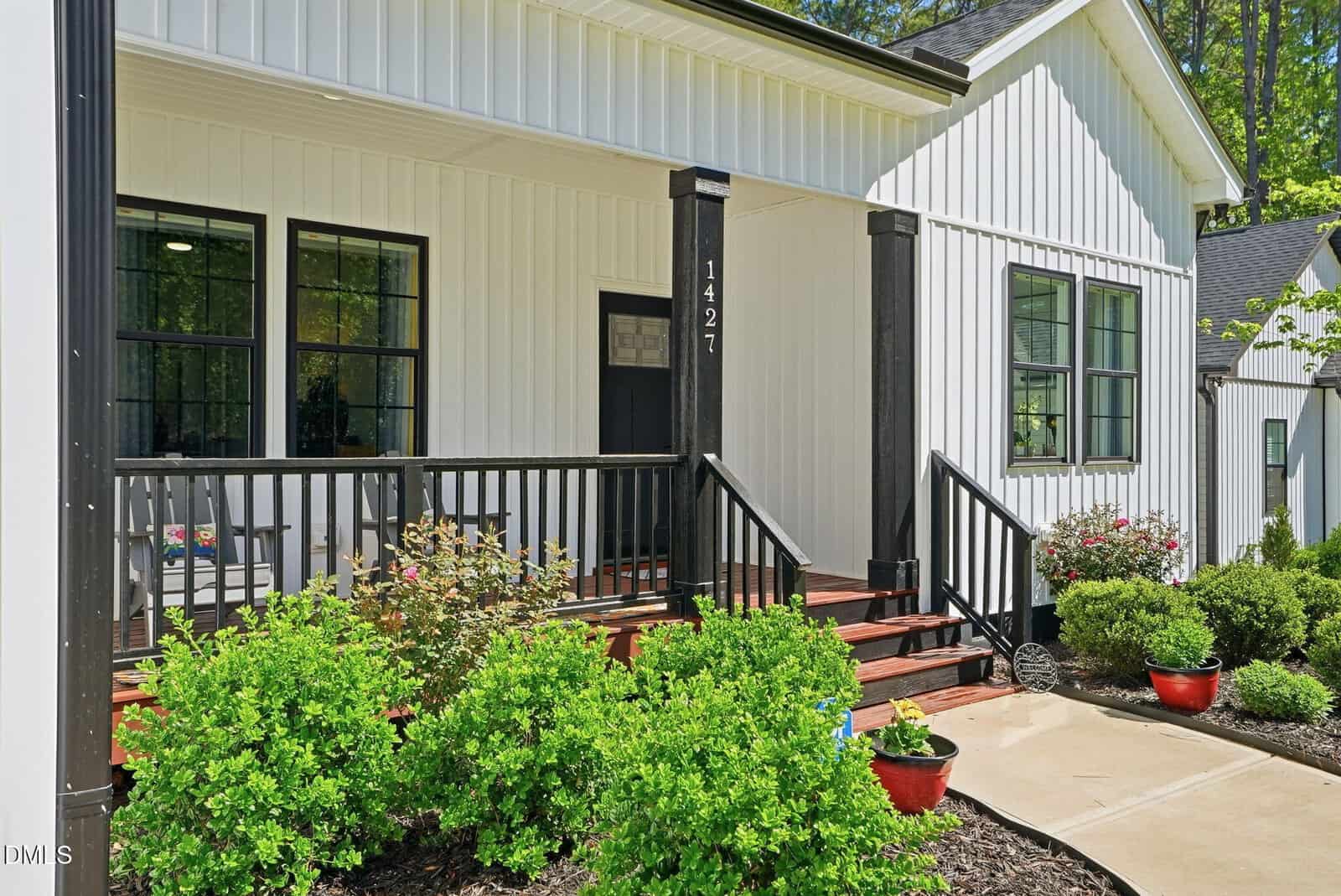 White house with black trim and porch railings, wooden steps, potted plants, shrubs, and a concrete walkway leading to the front door. The house number “1427” is displayed on a porch post.