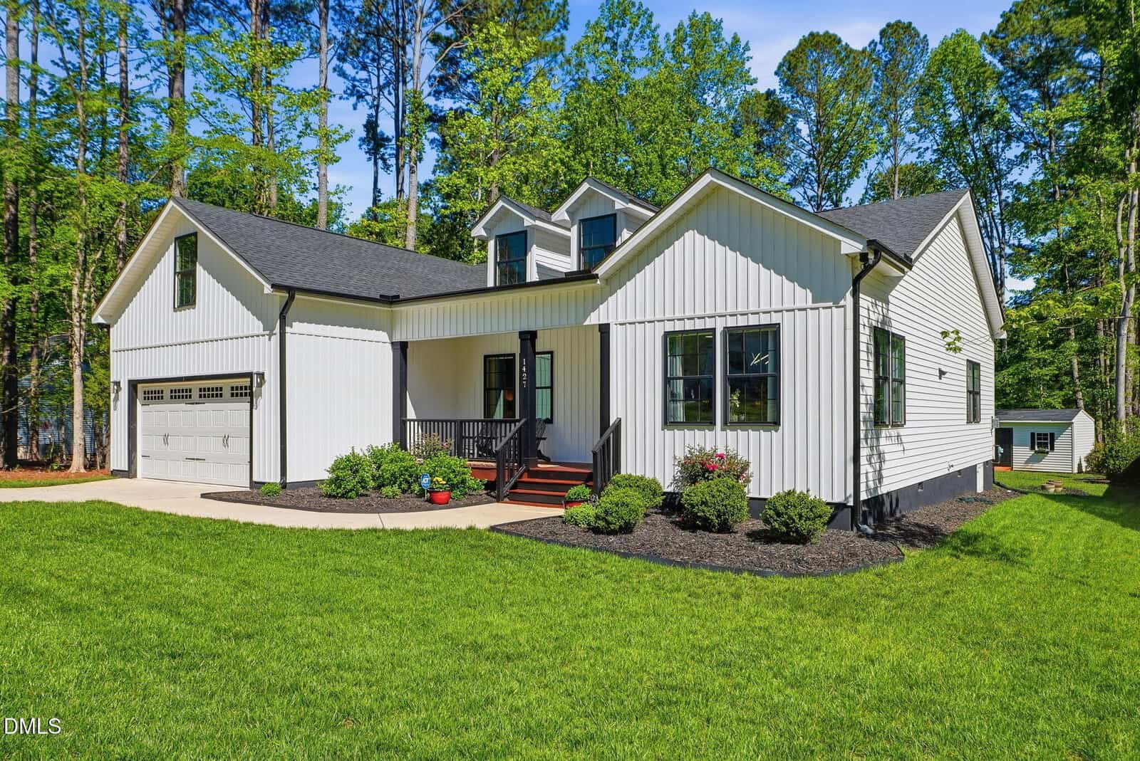 A modern white house with black trim, gabled roof, front porch, attached two-car garage, landscaped yard, shrubs, and green grass, surrounded by tall trees on a sunny day.