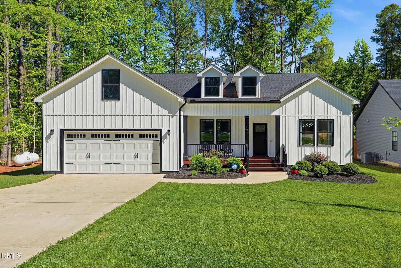 A modern white house with black trim, two dormer windows, and a two-car garage sits on a green lawn with landscaped bushes and flowers, surrounded by tall trees on a sunny day.
